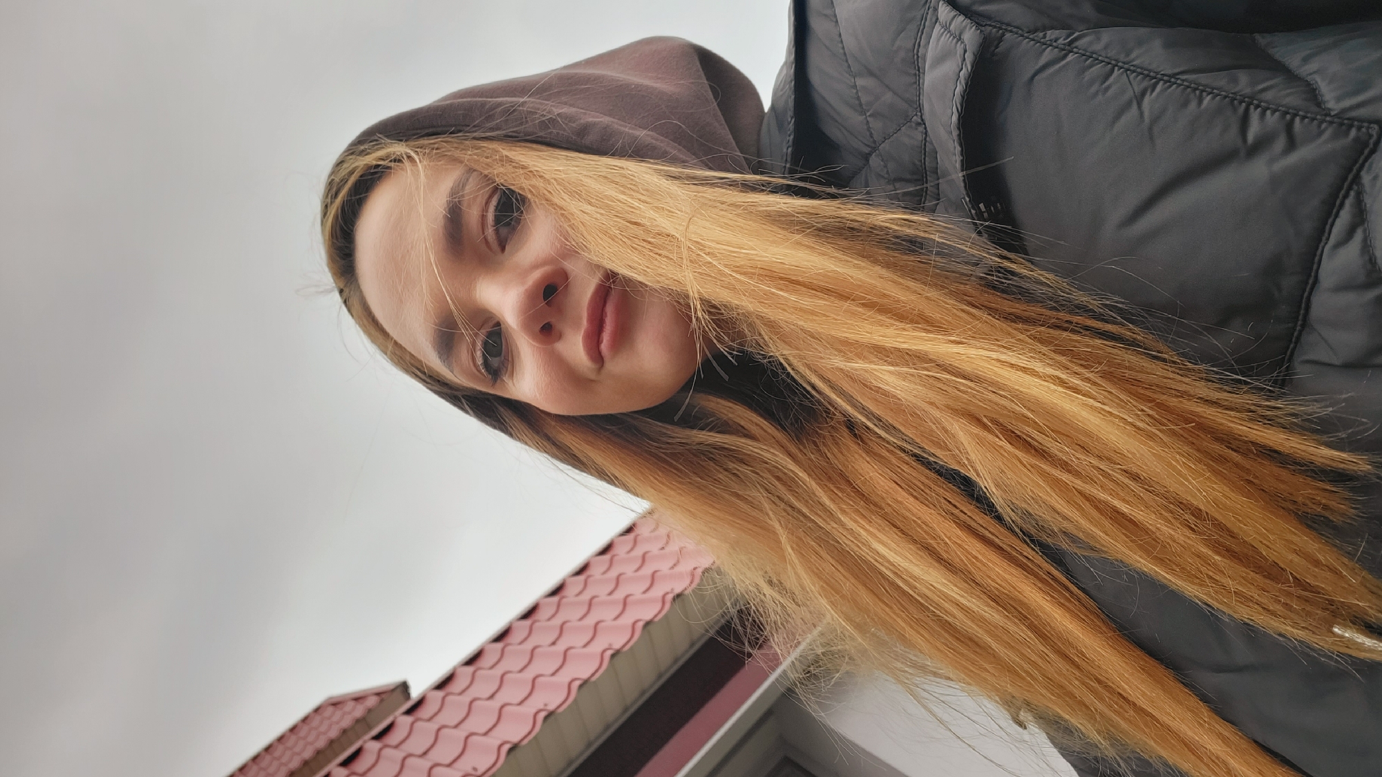 A woman with long, light-brown hair, wearing a dark-brown hooded sweatshirt and a black quilted jacket, looks down at the camera. The sky is overcast. In the bottom left corner is a building with a red roof. The image appears to be taken in the city of Chaikovsky, Russia.