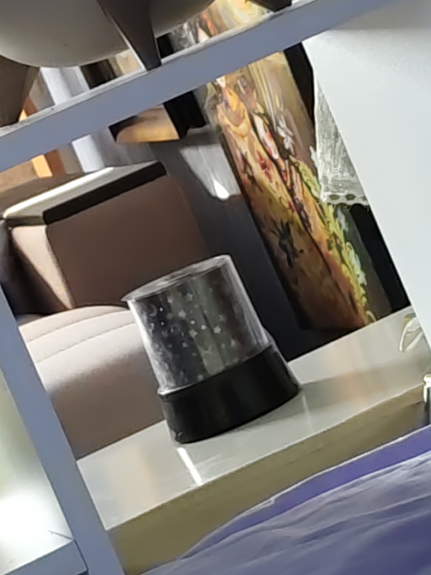 The image is an indoor shot, possibly a room or a studio, with shelves. On the shelf in the foreground, a cylindrical object with a black base and a clear, patterned top is visible. The patterned top appears to have star and crescent moon motifs. Behind this object, a large, light brown upholstered furniture item can be seen. Further in the background, a colorful painting or tapestry with floral elements is displayed on a dark surface. The overall lighting suggests an ambient indoor setting, with some reflections present on the surfaces. There is no information available to determine the location in the city of Rostov, Russia.