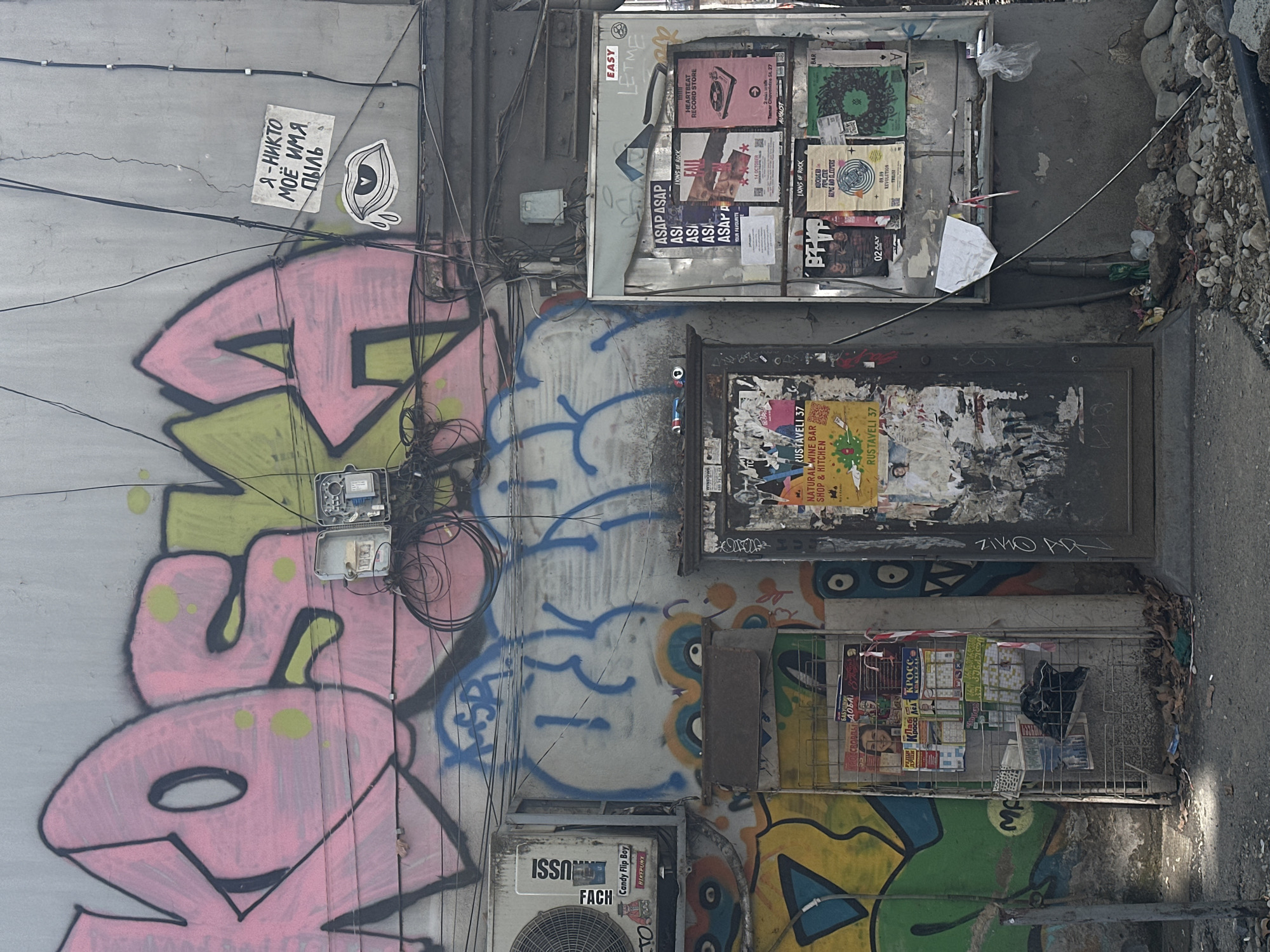 The image depicts a weathered concrete wall covered in graffiti and posters. The graffiti features large, stylized pink and green letters, along with other spray-painted designs in blue, yellow, and orange. Several posters, some faded and torn, are affixed to the wall, advertising events, bars, and shops. Among the visible posters are ones for "ASAP ASAP", "LIONS OF ROCK", and "HEARTBEAT RECORD STORE".
In the lower left of the frame, an air conditioning unit is mounted on the wall. To the right of the air conditioner is a metal rack filled with various publications, including magazines and what appear to be calendars. A dark plastic bag is visible within the rack.
Further to the right, a dark-colored metal door is set into the wall, also covered in peeling posters and graffiti. Above the door and extending across the wall are numerous black electrical wires and conduits, suggesting an urban setting with visible infrastructure. On the wall to the upper right, a small handwritten sign with Cyrillic text and an eye graphic sticker are present.
The ground in front of the wall is paved with asphalt and littered with small rocks, debris, and fallen leaves. The overall impression is one of urban decay and artistic expression, characteristic of many city backstreets. While the specific location is not explicitly stated in the image, the presence of Cyrillic script on a sign suggests a region where it is used. Based on the style of graffiti and the overall aesthetic, this scene is consistent with locations in Tbilisi, Georgia, which often feature such urban art and exposed infrastructure.