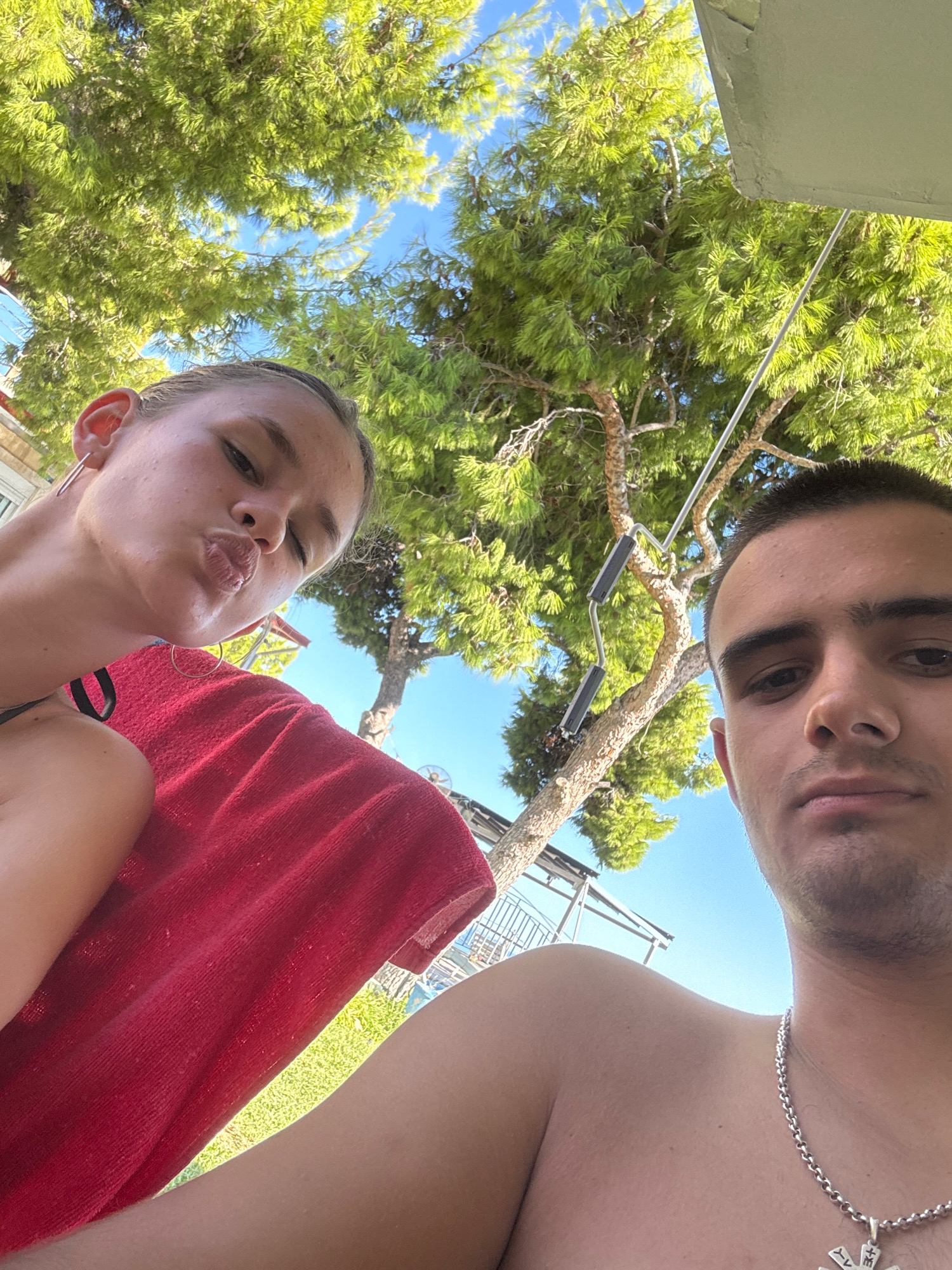 The image shows a close-up, high-angle selfie of two young adults, a female and a male, outdoors under a canopy of green trees with a clear blue sky visible. The female, positioned on the left, is looking at the camera with her lips puckered and wearing large hoop earrings and a dark-colored swimsuit strap. A vibrant red towel is draped over her shoulder and chest. The male is on the right, looking directly at the camera with a neutral expression. He is wearing a silver chain necklace with a pendant. The background consists of lush green foliage from trees, with some sunlight filtering through. In the distance, there are glimpses of what appears to be a railing, possibly from a balcony or a beachside structure, and bright green grass. The overall setting suggests a warm, sunny outdoor environment, potentially a vacation spot. The composition, with the subjects framing the shot from a low perspective looking up at the trees, is characteristic of a selfie taken in a relaxed, outdoor setting. It is not possible to determine the specific location within the Municipal Unit of Megara, Greece, based solely on the visual information provided in the image.