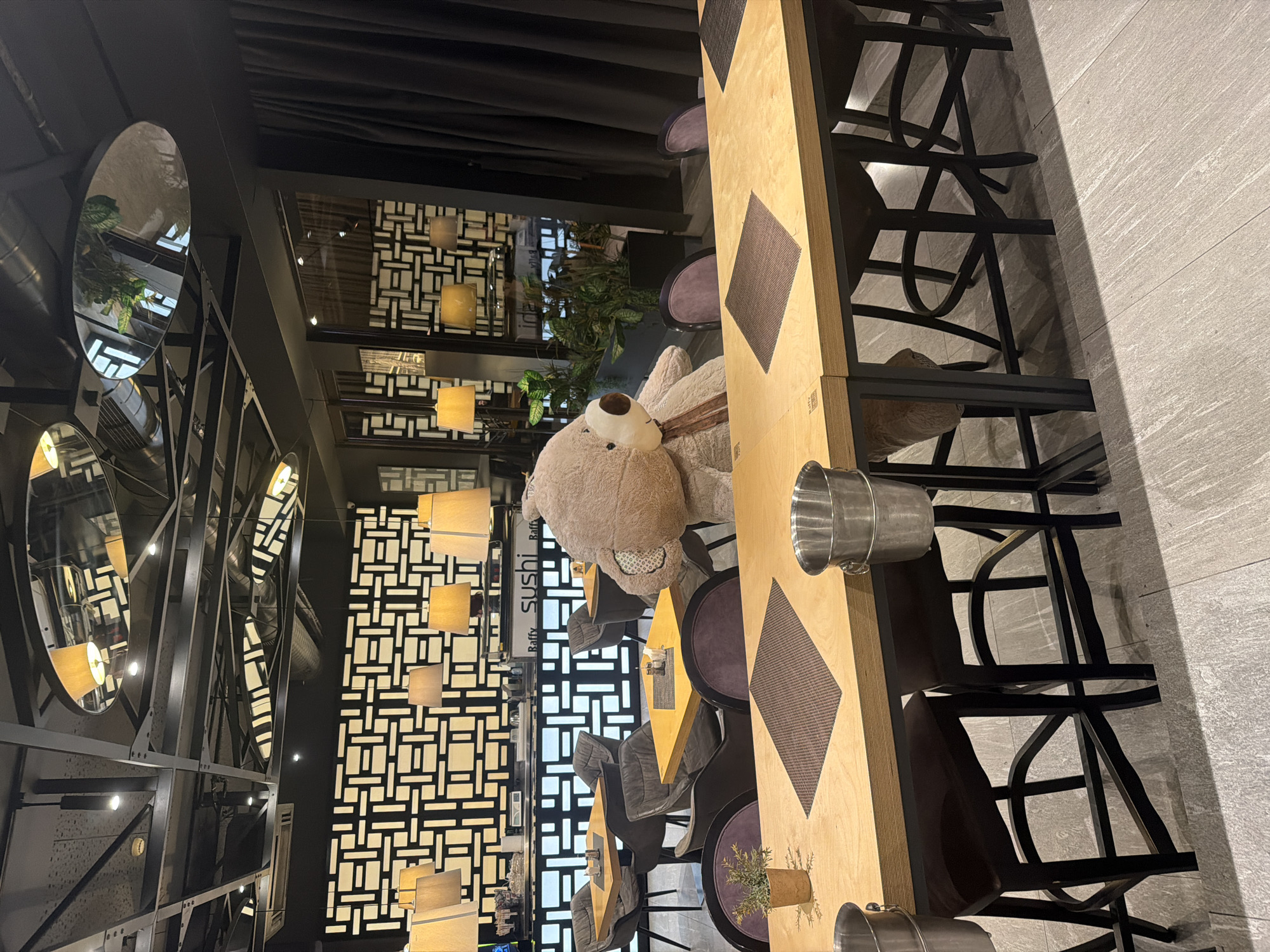 The image shows the interior of a restaurant or bar, featuring a long wooden table with chairs. A large teddy bear sits at the table. The restaurant has a modern design with decorative mirrors and geometric patterns on the walls. The location is likely a restaurant in the city of Plovdiv, Bulgaria, based on the signage “Raffy sushi”.