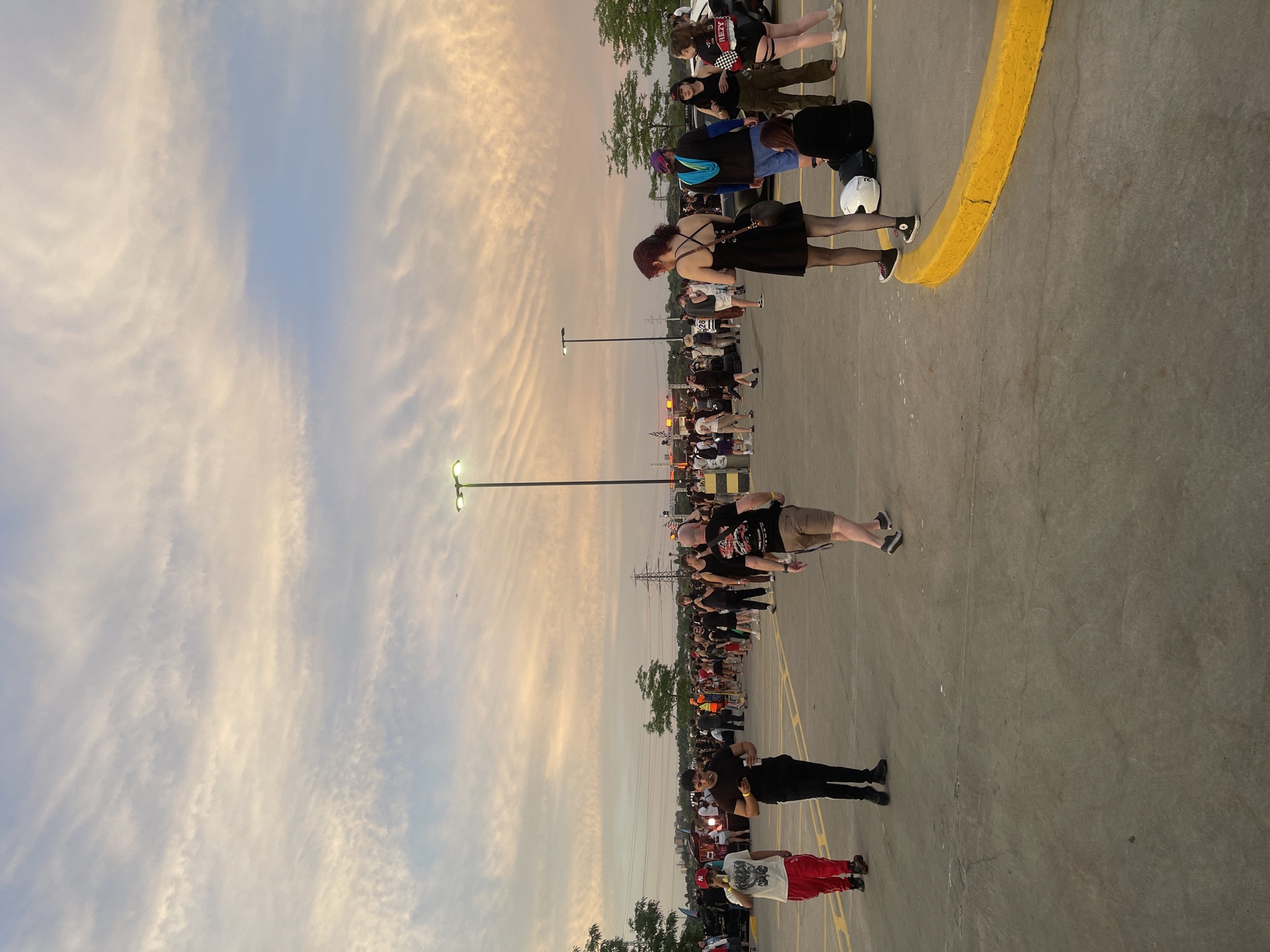 The image depicts an outdoor gathering of people in a parking lot at dusk. The sky is filled with streaky, wispy clouds in shades of orange, pink, and blue, suggesting a sunset or sunrise. Several streetlights are visible, illuminating the scene.
In the foreground, people are walking and standing. A man in a red hat and red pants is on the left. To his right, a man in a black t-shirt and black pants is smoking. Further to the right, a man in a black t-shirt with a graphic design on the back is walking away from the camera. A woman in a black dress and fishnet stockings is also visible, as is another woman in a black top and green cargo pants, and a woman in a black and white top and shorts. Several other individuals are scattered throughout the scene, some appearing to be interacting with each other.
In the background, more people are gathered, and there appear to be some temporary structures or booths, possibly for an event. Trees and power lines are also visible in the distance. The ground is paved asphalt, with yellow painted lines marking parking spots or traffic lanes.
The specific location in the city of Toronto, Canada, cannot be determined from the visual information provided in the image.