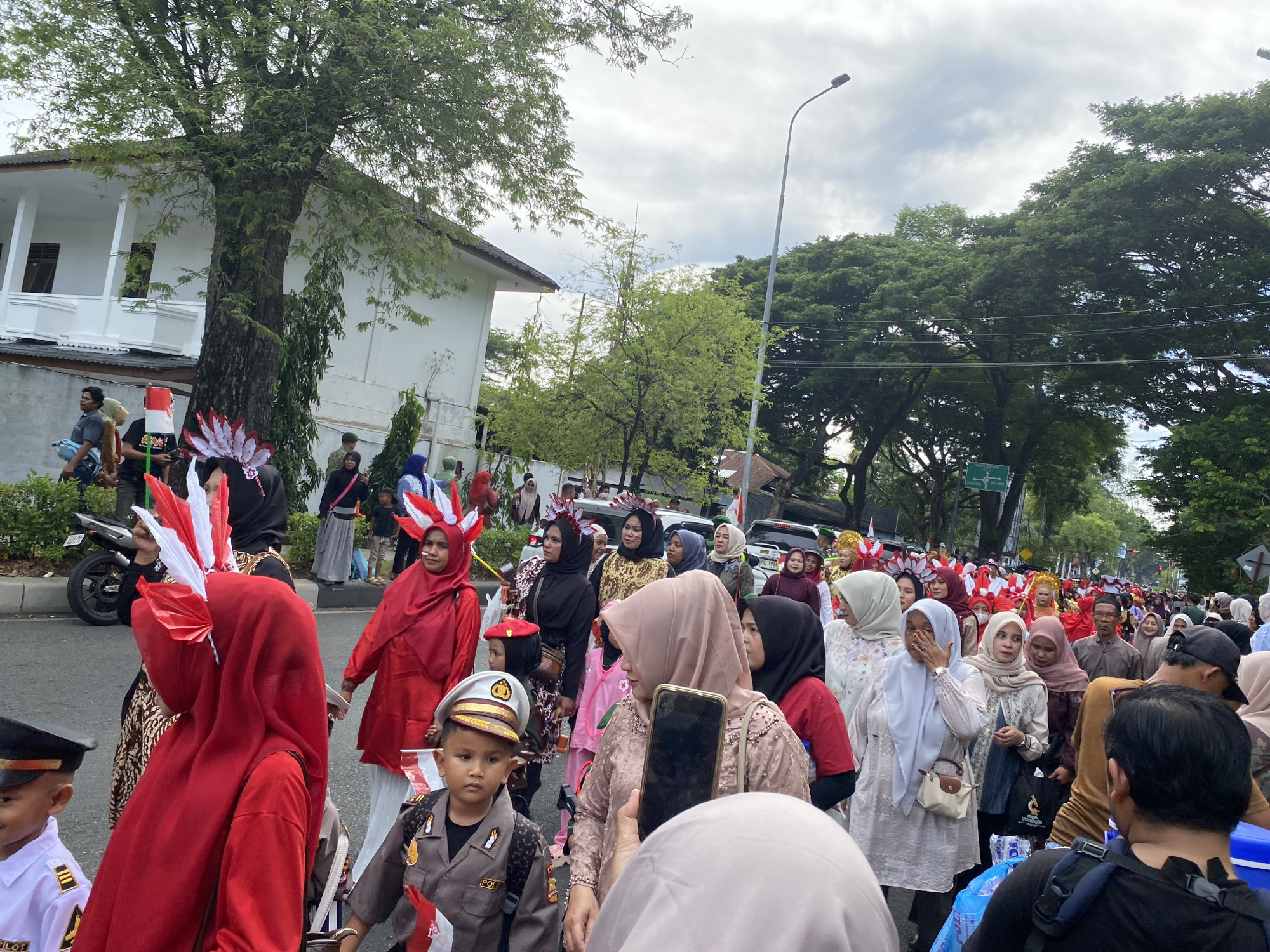 The image shows a street scene in Banda Aceh, Indonesia, where a parade is taking place. A large crowd of people, predominantly women, are walking on a paved street lined with trees and buildings. Many participants are wearing traditional Indonesian attire, including colorful hijabs and dresses. Some individuals are adorned with festive headdresses resembling feathers or flags, predominantly in red and white, the colors of the Indonesian flag.
In the foreground, a young boy dressed in a police uniform, complete with a hat and badge, walks with a serious expression. To his left, a person in a bright red hijab with a white and red feathered headdress is prominent. In the background, more people are visible, some carrying small Indonesian flags. Vehicles, including a motorcycle and cars, are also present on the street, suggesting it is a public event. The sky is overcast with a few clouds. The architecture of the buildings suggests an urban setting. The overall atmosphere appears to be celebratory and communal.