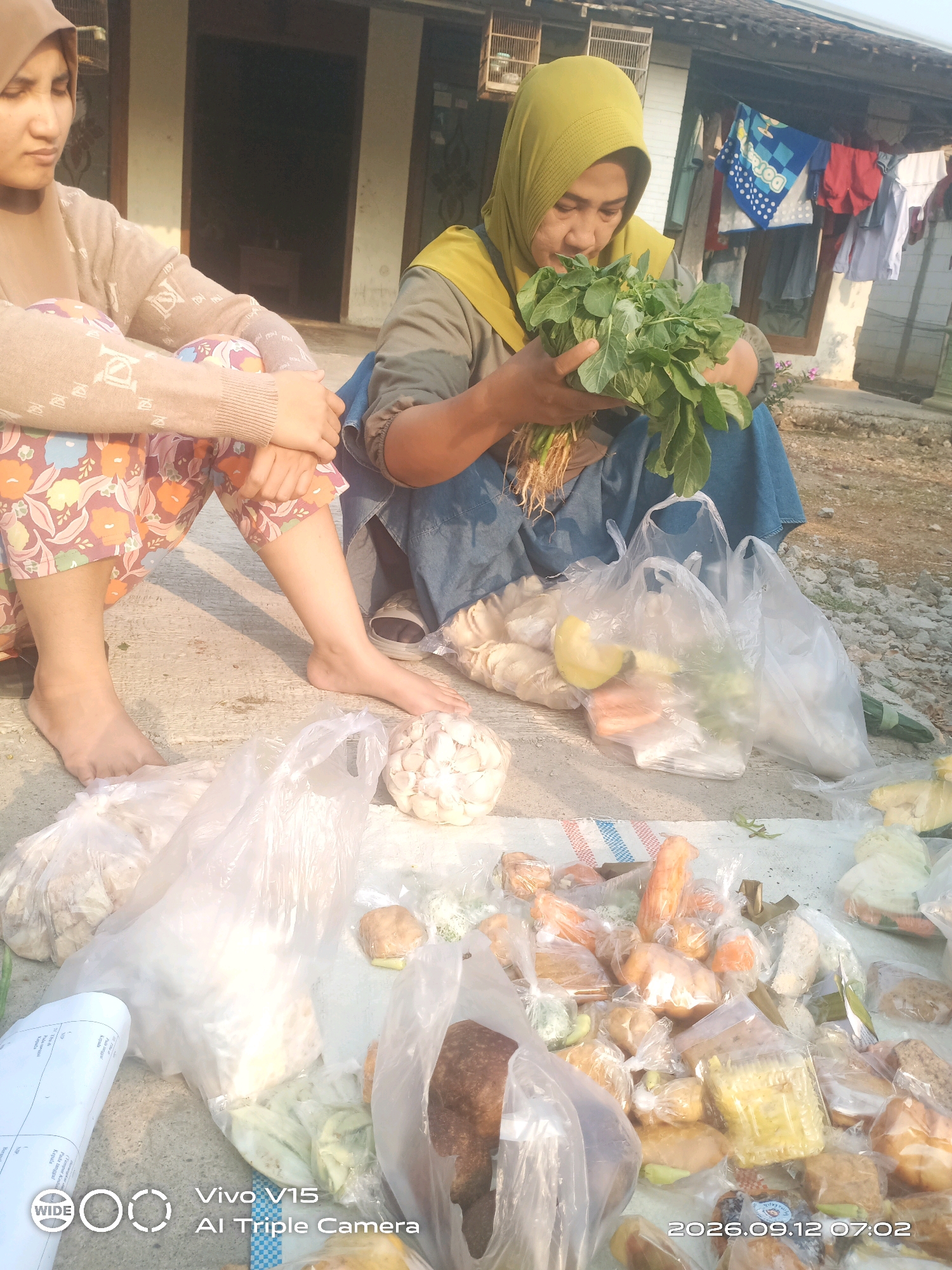 The image depicts an outdoor scene, likely in Plosorejo, Indonesia, where two individuals are seated on a concrete surface, surrounded by various food items. On the left, a person wearing a patterned skirt and a light-colored cardigan is shown from the waist down, their legs crossed. To the right, a woman wearing a yellow hijab and a blue skirt is holding a bunch of leafy green vegetables, seemingly examining them. She is also seated on the ground. The ground is covered with a white tarp, on which numerous plastic bags filled with food are arranged. These food items include what appear to be fried snacks, bread, vegetables like carrots and chilies, and a head of garlic. In the background, a simple building with a tiled roof is visible, with laundry hanging on a clothesline. The lighting suggests it is daytime. The overall impression is of a local market or a personal sale of food items in a semi-urban or rural setting.