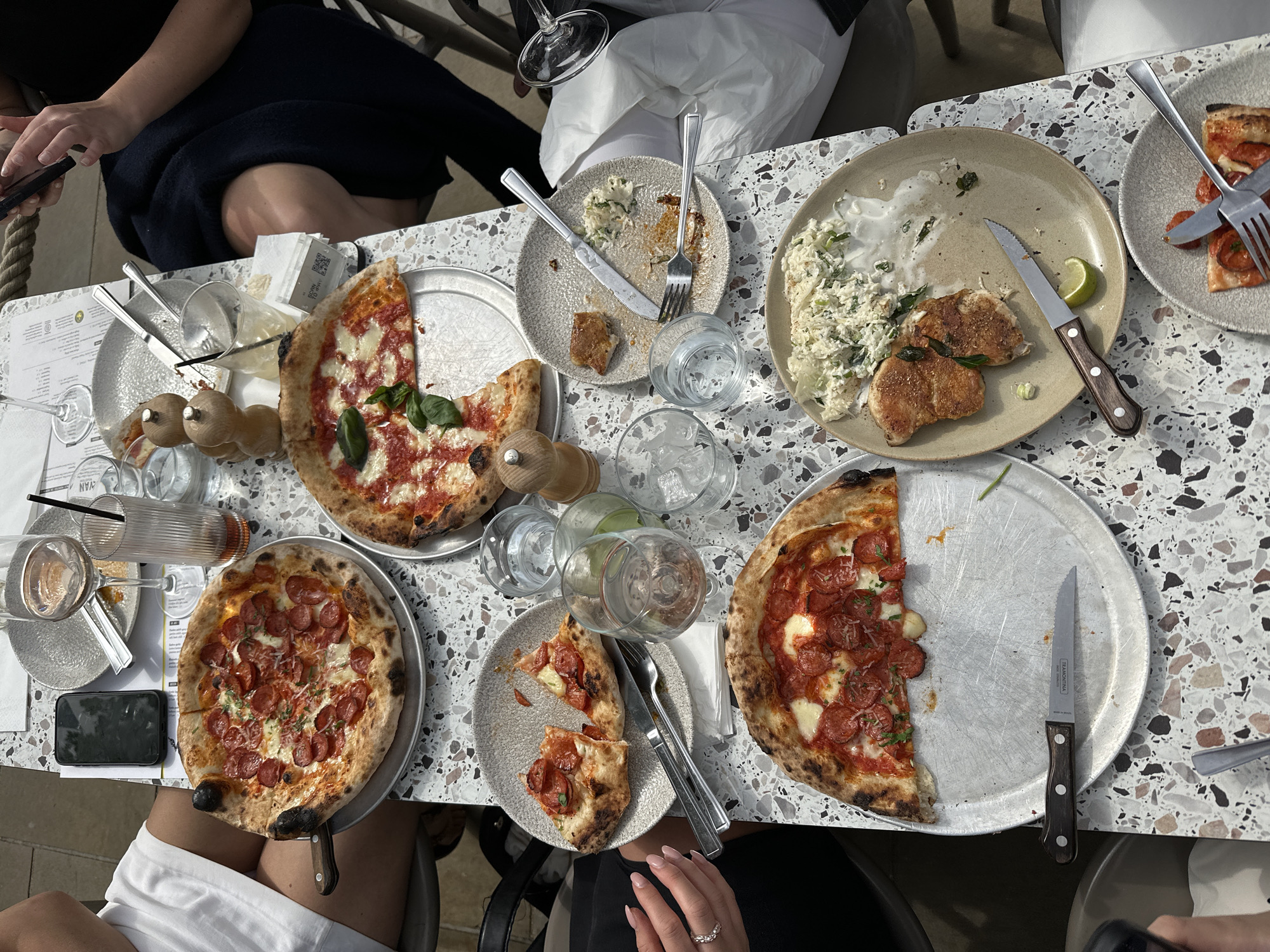 Captured from an overhead perspective, the scene portrays a bustling table setting in what appears to be an outdoor dining area. The table, marked with a speckled pattern, is laden with food and drink. Three pizzas, one with what looks like pepperoni and two with a red sauce base, sit on metallic serving trays. Plates hold the remnants of food, including a dish containing meat and a creamy, shredded slaw-like substance. Utensils such as knives and forks are scattered among the plates. Several glasses, some filled with liquid, add to the sense of activity. Partial views of people seated around the table are visible, including glimpses of clothing, hands, and accessories like a ring. A mobile phone rests beside one of the pizza trays. The setting lacks distinct identifiers to determine its precise location in the city of Unknown, Unknown.