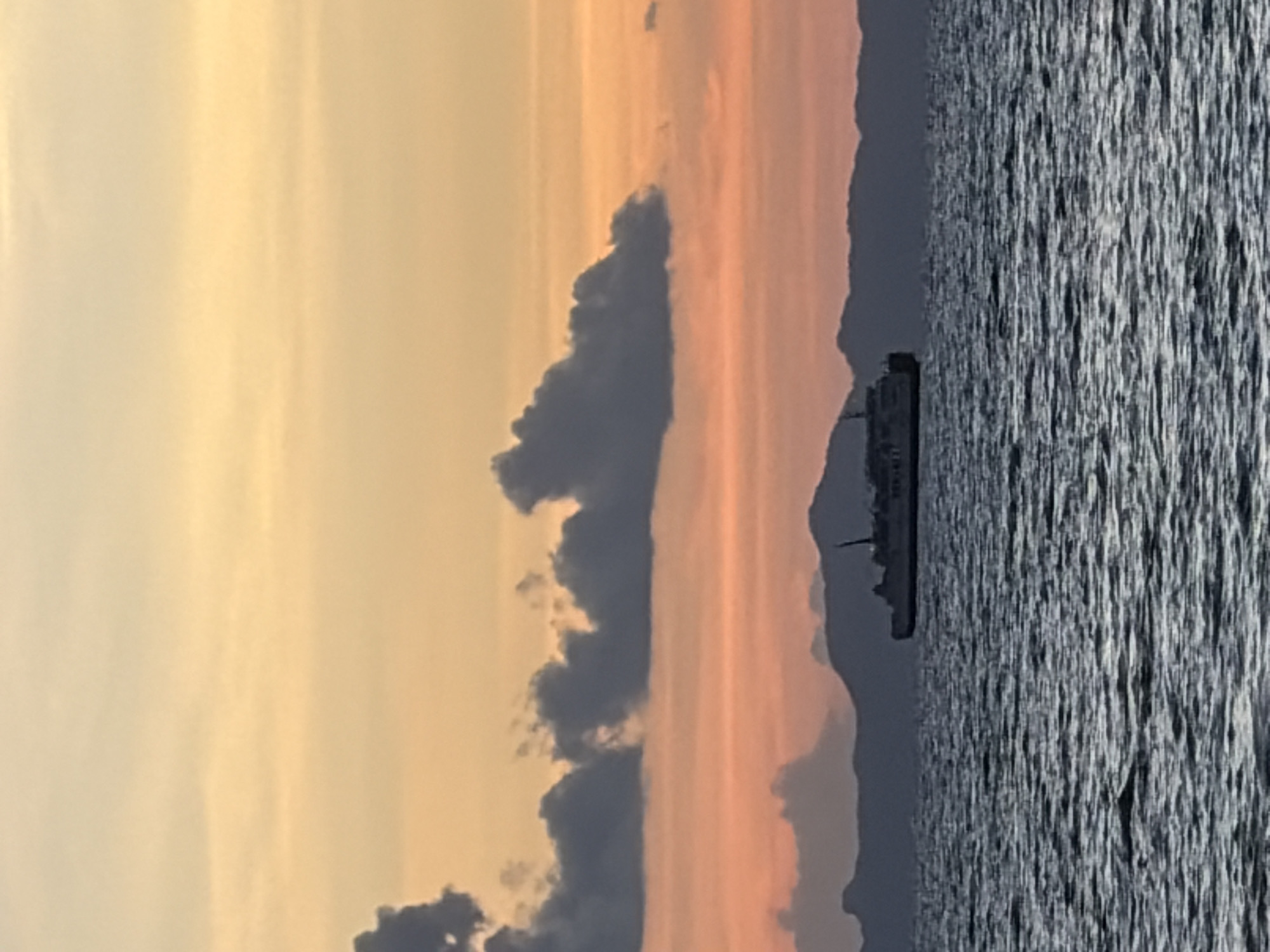 At sunset in Baan Taling Ngam, Thailand, a ferry boat travels across the ocean. The sky is a blend of orange and yellow hues, with dark clouds scattered across it. In the distance, a mountain range silhouettes against the vibrant sky. The ocean water is dark and rippled, reflecting the colors of the sunset. The ferry is a large vessel with multiple decks and a single smokestack. It is positioned in the middle of the frame, with the mountains and sky behind it. The overall scene is one of tranquility and natural beauty.