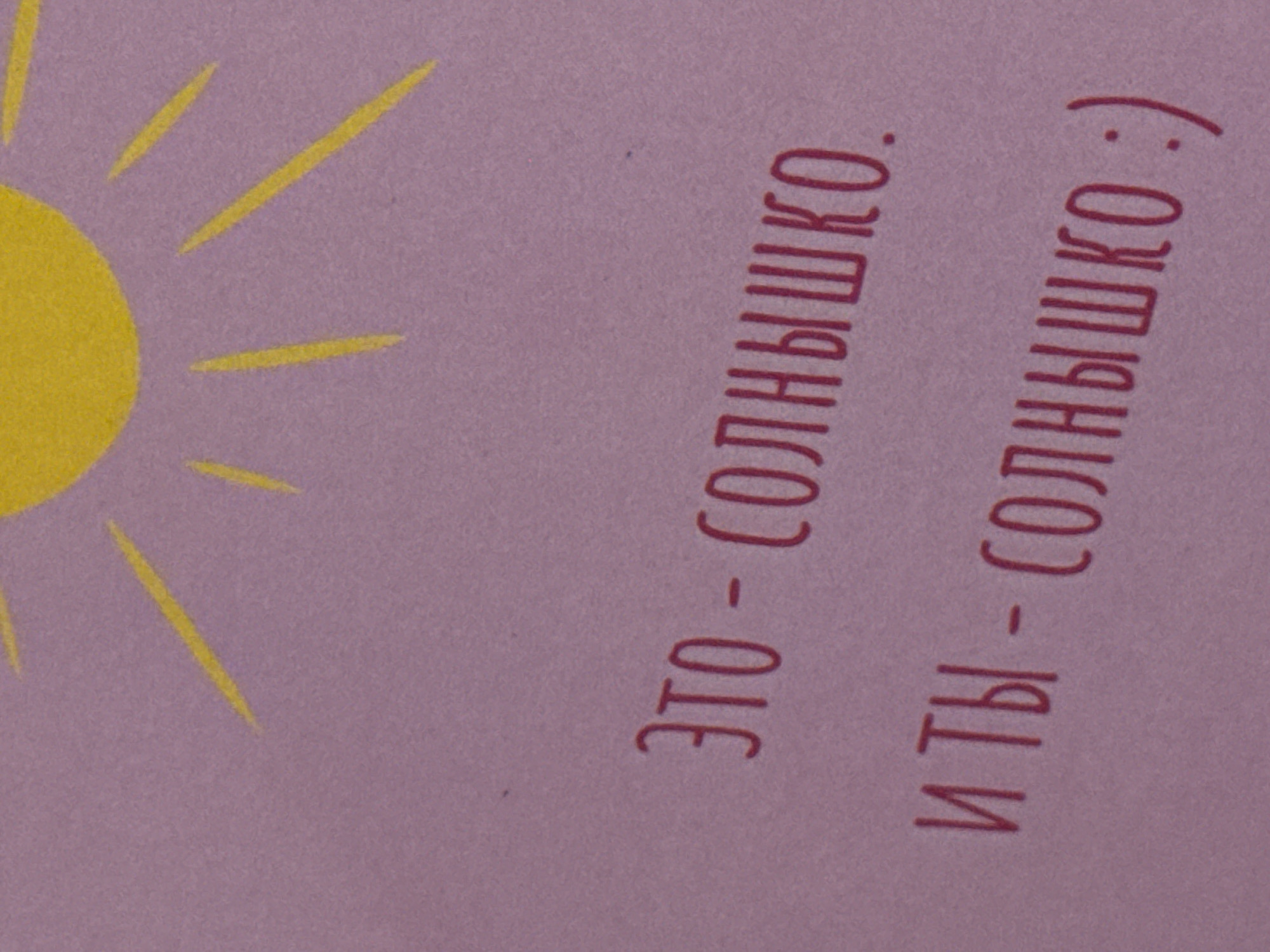 The image shows a pale purple background with a yellow sun graphic in the upper left corner. To the right of the sun are two lines of text in Russian, written in dark red. The first line reads "ЭТО - СОЛНЫШКО." and the second line reads "И ТЫ - СОЛНЫШКО :)". The text translates to "This is the sun." and "And you are the sun :)". There are no people or other objects in the image. The location is not ascertainable from the provided image.