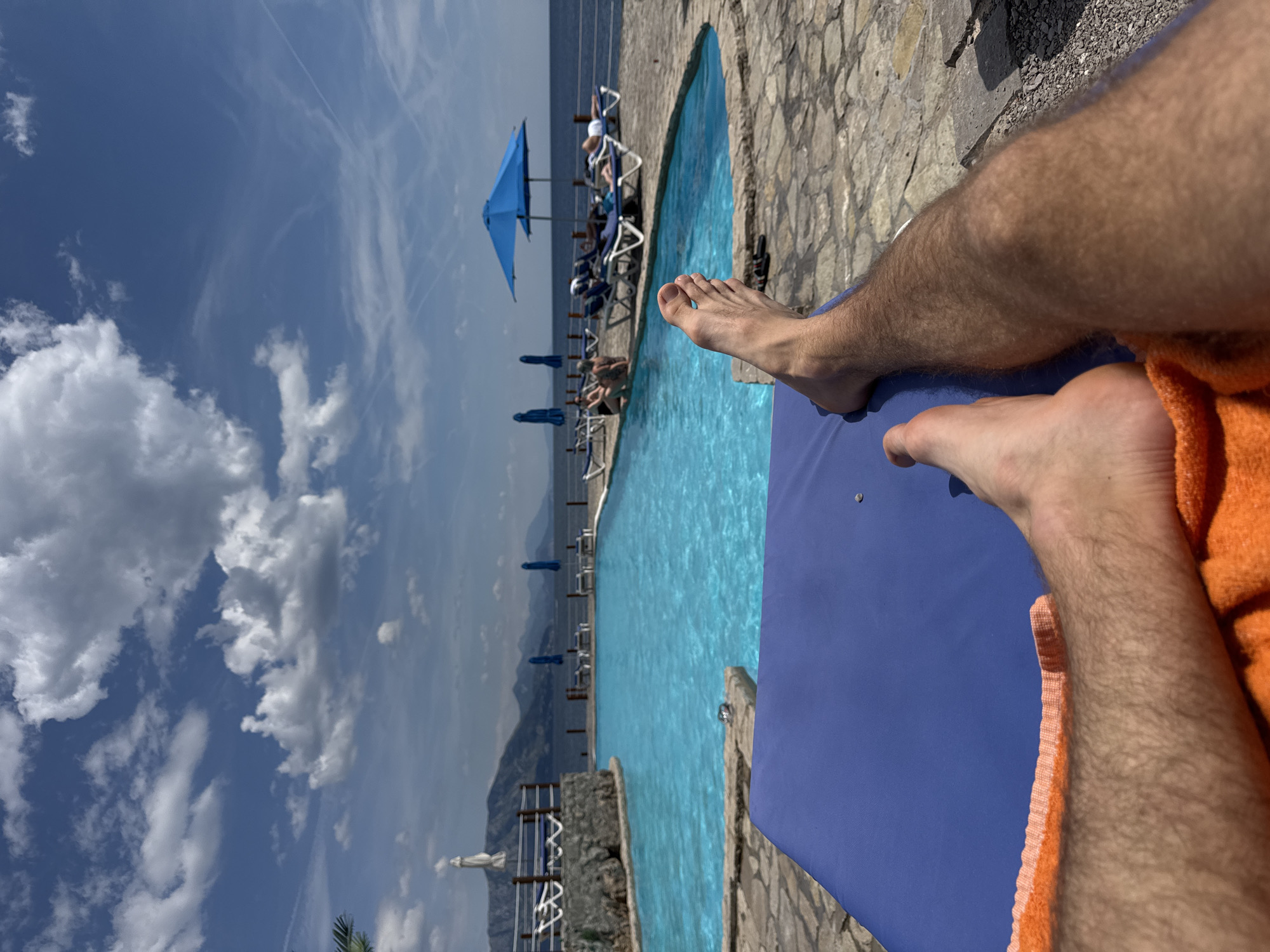 The image depicts a person relaxing on a blue lounge chair by a pool. The person's hairy legs and feet are visible in the foreground, with an orange towel partially obscuring one leg. The turquoise pool stretches out towards the horizon, lined with blue sun umbrellas and lounge chairs. Several people are relaxing on the lounge chairs near the pool's edge. The background features a hazy mountain range and a partly cloudy blue sky. The pool is surrounded by a stone border. The scene suggests a resort or vacation setting in Prijevor, Montenegro, a town not found in Montenegro. The location is likely in the larger region of Prijevor, which is in Montenegro.