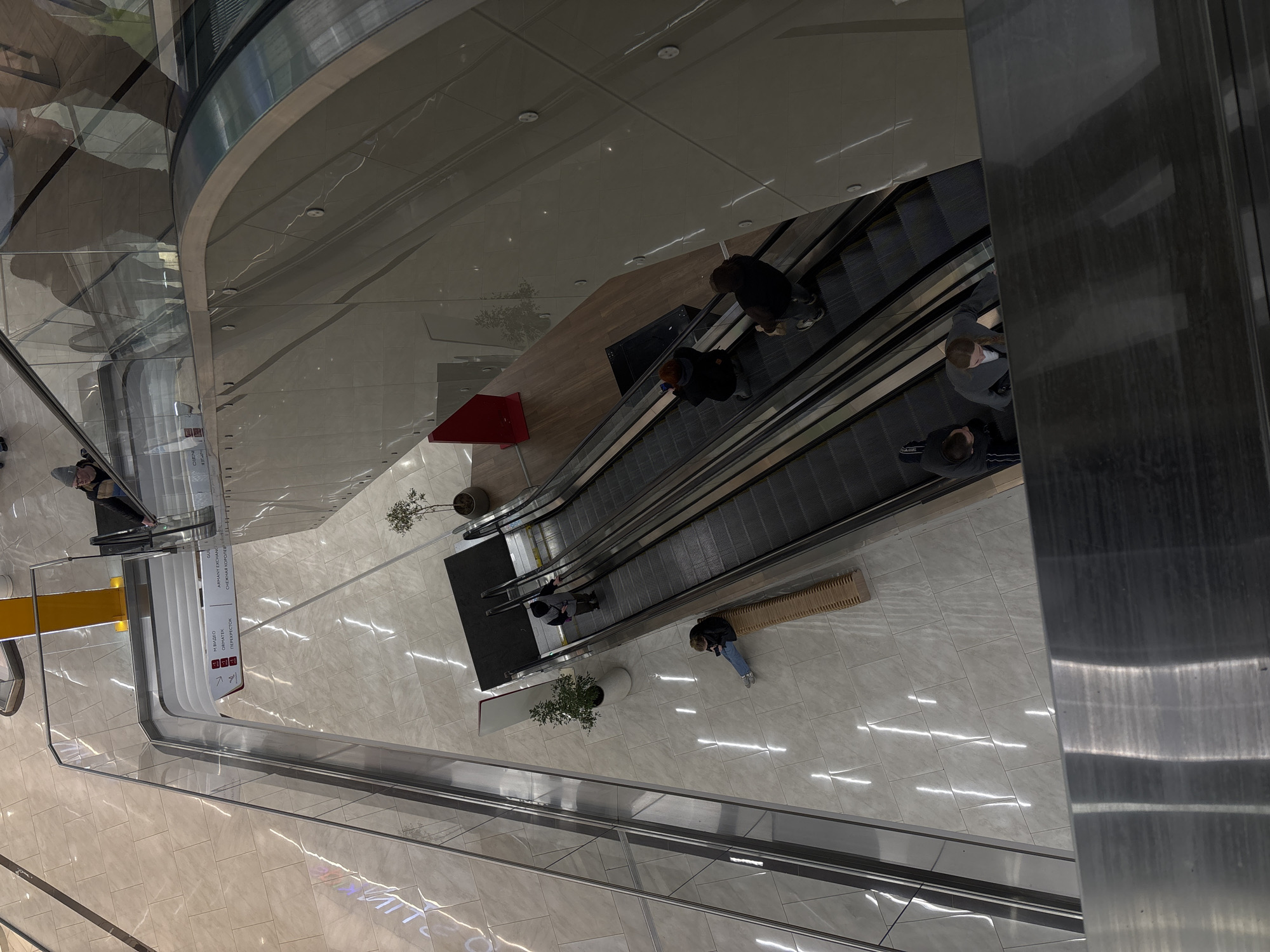 This high-angle, top-down image captures the interior of a modern, multi-level building, likely a shopping mall, located in Perm, Russia. The scene is dominated by a pair of escalators serving as a central vertical transport system, bordered by transparent glass balustrades and polished, light-toned tiled flooring. Various reflections from overhead lighting and architectural elements create intricate patterns on the shiny surfaces.
Several individuals are present, engaged in typical mall activities. A few people are seen riding the escalators, either ascending or descending. On the lower visible level, one person is seated on a long, light-colored wooden bench, while another stands near a glass railing. Small potted plants are strategically placed around the area, adding a touch of nature. A distinctive bright red, triangular object rests on the floor near the escalator base.
A vertical white information sign is prominently displayed, listing various stores and services. Visible text on the sign includes "М.ВИДЕО" (M.Video, an electronics retailer), "ОПЕРЕСТОК" (likely Perekrestok, a supermarket chain), "АРМАНИ ЭКСПРЕСС" (Armani Exchange), "СНЕЖНАЯ КОРОЛЕВА" (Snezhnaya Koroleva, a fur and fashion store), and "GALY". The sign also features directional arrows and floor numbers like '2' and '3'. The overall ambiance is bright and clean, typical of a well-maintained modern commercial space.