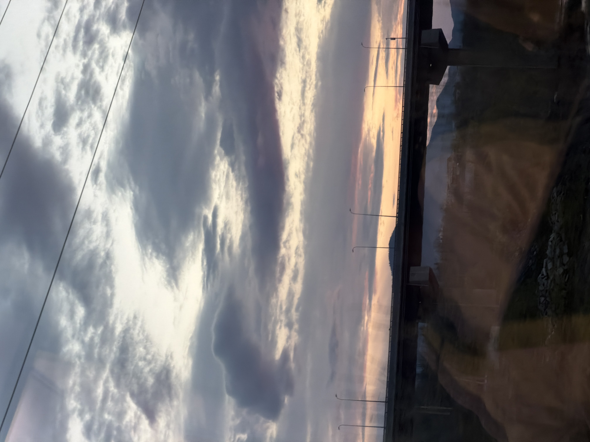 The image shows a cloudy sky with a hint of sunset, likely in Japarauli, Georgia, as indicated by the request. A bridge with streetlights spans the view. A mountain is in the distance. A reflection is visible on the right side of the image. Power lines stretch across the sky.