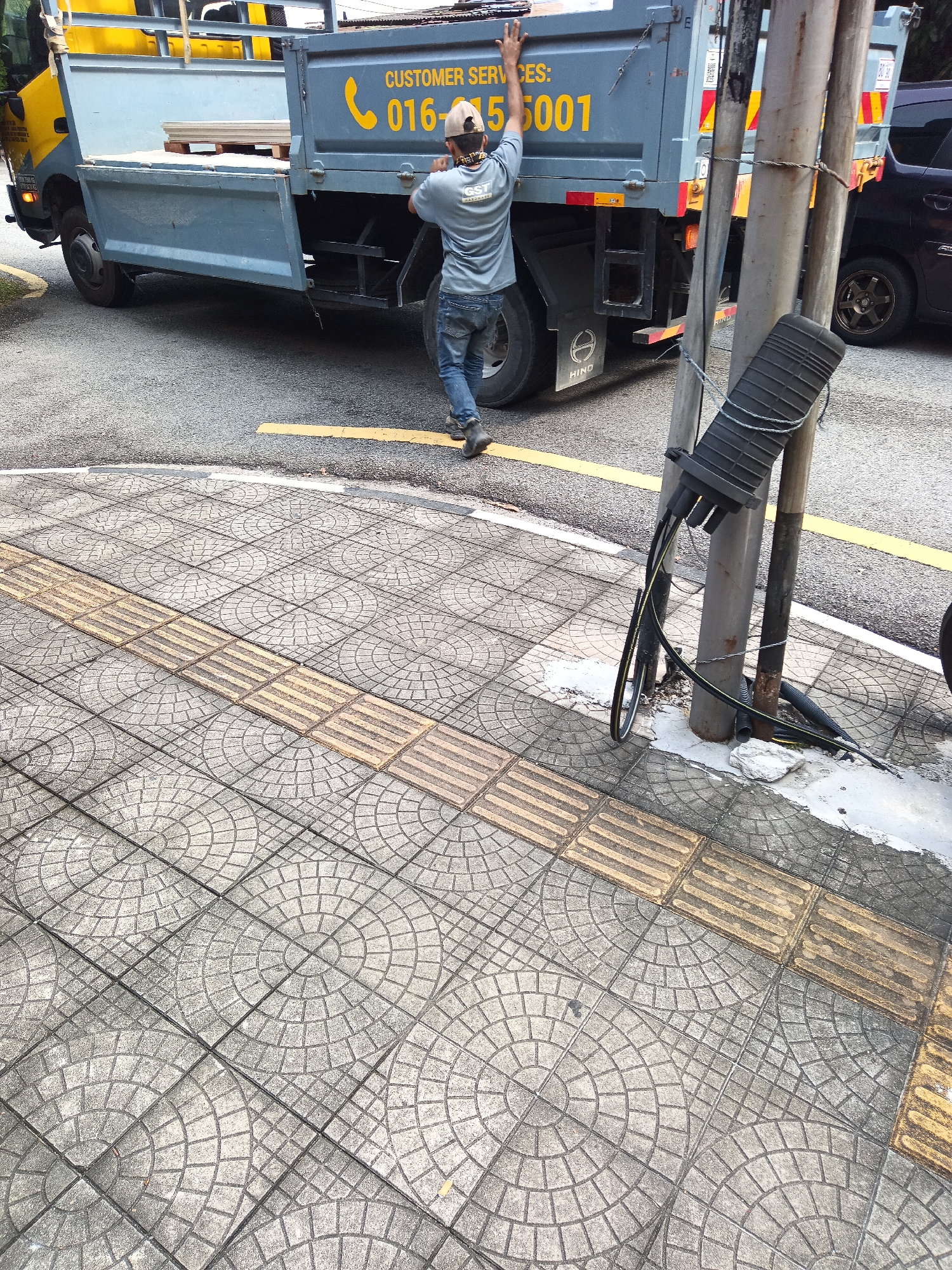 A man in a gray t-shirt and blue jeans stands next to a grey truck with yellow lettering that says "CUSTOMER SERVICES: 016-55001". He is pushing the back of the truck with his hands. The truck is parked on a street with yellow lines. In the foreground, there is a sidewalk with grey circular patterned tiles and tactile paving. A utility pole with cables attached is also visible. The scene appears to be in Kuala Lumpur, Malaysia, given the context of the truck's signage.