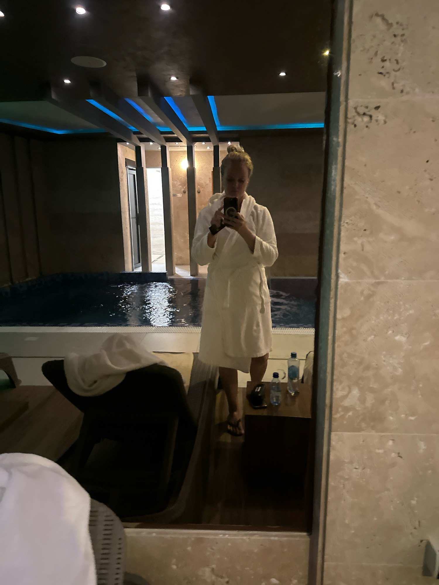 A woman in a white robe takes a selfie in a dimly lit indoor pool area. The pool is dark blue and reflects the ceiling lights, which are modern and have blue accents. The woman is standing to the right of the pool, and in front of her is a small table with two water bottles and a phone. To the left of the pool is a lounge chair with a white towel draped over it. The walls are made of textured stone, and there is a large mirror reflecting the scene. The overall impression is one of luxury and relaxation. The location is a spa or hotel pool area in Belgrade, Serbia.