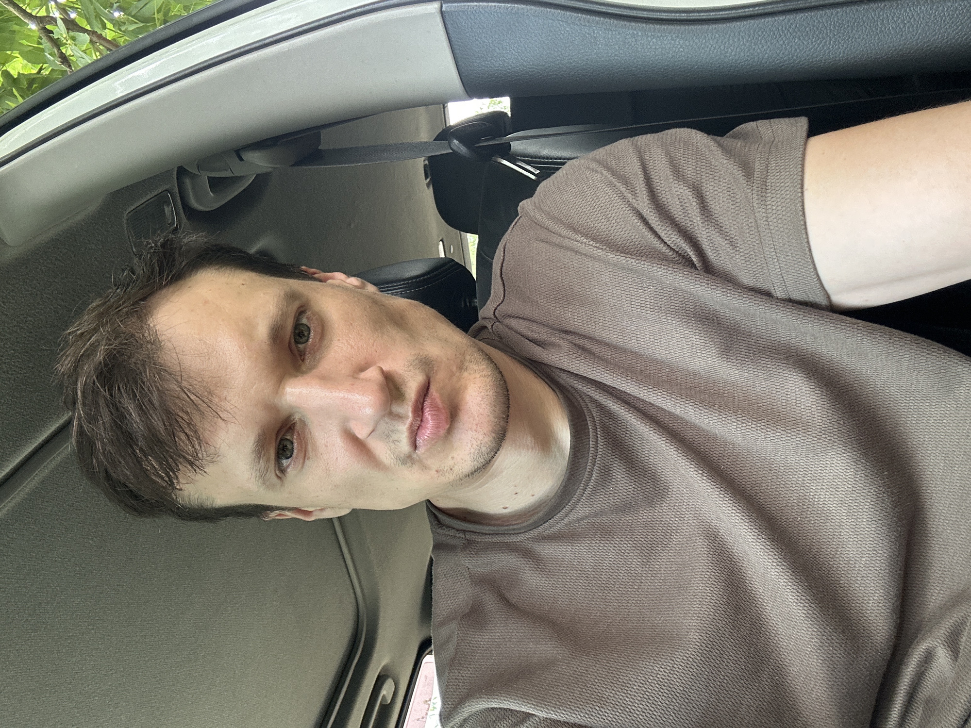 A close-up, waist-up, selfie-style photograph shows a man sitting in the driver's seat of a vehicle. The man has light skin, brown hair, and green eyes. He is wearing a brown t-shirt. The interior of the car is visible, including a sunroof, a seatbelt, and part of a headrest. Green foliage is visible through the car window, suggesting the vehicle is outdoors in a vegetated area. The photograph was taken on June 24, 2023, at 12:33 PM in Makiv, Ukraine.