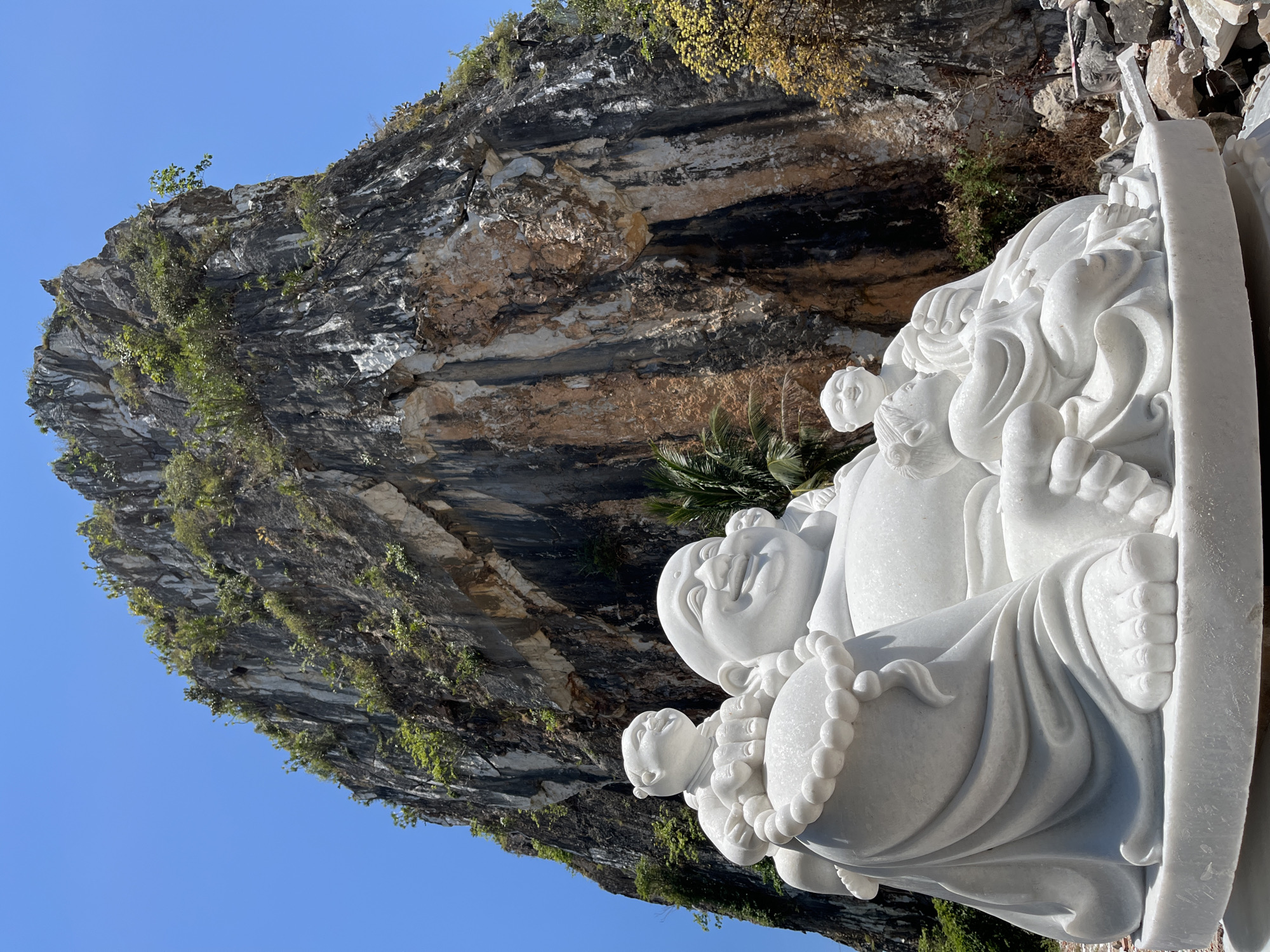 The image shows a white marble statue of a Budai (Laughing Buddha) figure with several children. The statue stands in front of a limestone cliff face covered in vegetation, with a blue sky in the background. The location is likely in the Marble Mountains (Ngũ Hành Sơn) in Đà Nẵng, Vietnam, known for their marble sculptures and Buddhist temples.