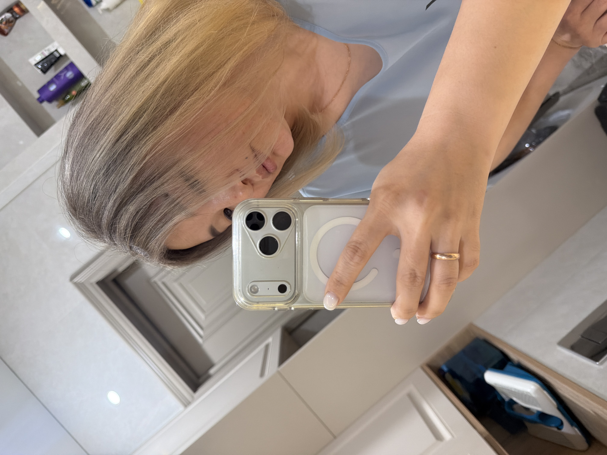 A woman is taking a mirror selfie. She is holding a phone in her right hand, which is in the foreground. She is wearing a light blue top and a gold ring on her right ring finger. Her nails are painted white. Her hair is blonde and styled in loose waves. Her face is partially obscured by her hair and the phone. In the background, there are shelves with various bottles and products, and what appears to be a bathroom or dressing area. The image does not contain any information that would indicate the location is the city of Astana, Kazakhstan.