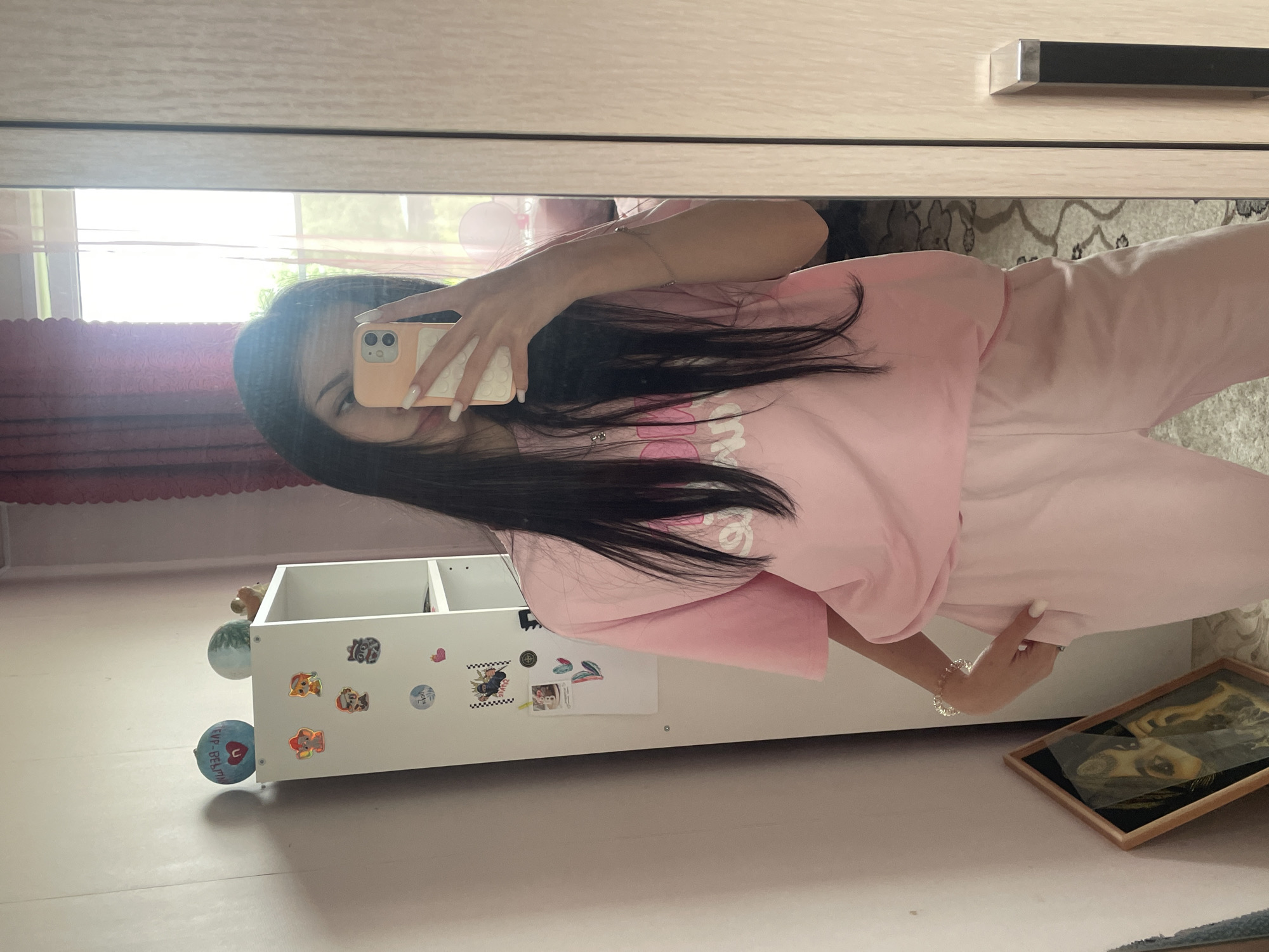 A young woman is captured in a mirror selfie, reflecting an indoor setting, likely a bedroom or personal space, bathed in bright daylight. The image is oriented vertically, showing the woman's reflection facing towards the viewer.
The subject is a young woman with long, dark hair, wearing a casual light pink long-sleeved top and matching light pink sweatpants. She holds a peach-colored smartphone with a white textured case in her left hand, positioned to take the picture. Her fingernails are painted white, and a delicate bracelet adorns her right wrist.
In the background, visible through the mirror's reflection, is a light-toned wooden wardrobe or closet with a modern black and silver handle. A window with red curtains or blinds is partially visible, showing bright light and greenery outside.
On the floor, a white open shelving unit, possibly on wheels, is decorated with various stickers, including Paw Patrol characters, a heart, and a checkerboard pattern. Attached to the side of the unit are two spherical objects, one blue and one green. The blue sphere features Cyrillic text "ГУР-ВЕРИТ" and a heart symbol. Leaning against the wall on the right side of the frame is a wooden-framed picture or artwork, depicting what appears to be a stylized face or animal. A light-colored, patterned rug is partially visible on the floor.