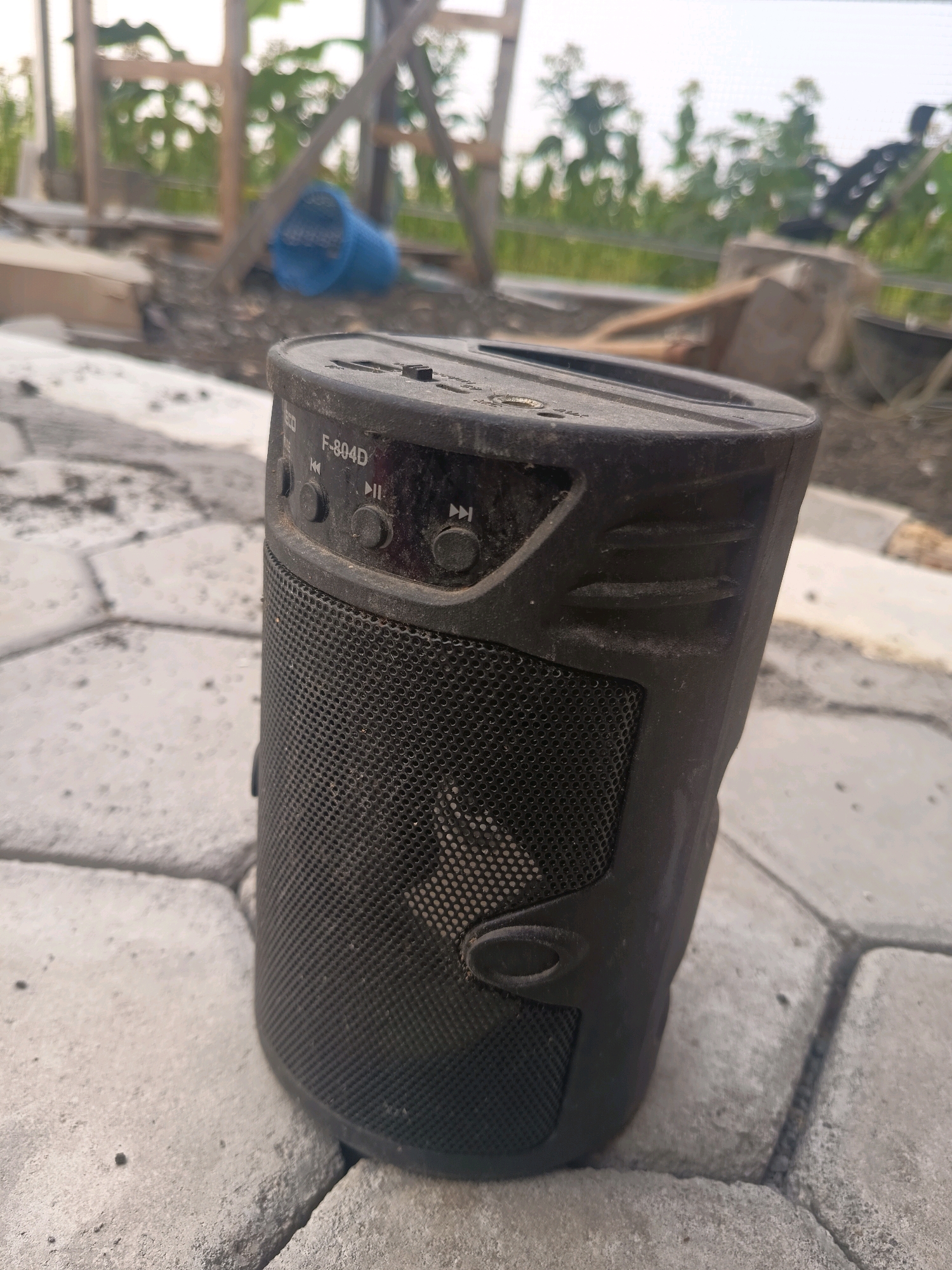 A black, cylindrical Bluetooth speaker rests on a light gray, textured paver surface. The speaker is dusty and shows signs of wear. On the top surface of the speaker, a model number "F-804D" is visible, along with several control buttons, including play/pause and track skip icons. A black mesh covers the front of the speaker. The background is blurred but appears to be an outdoor area with some wooden structures and greenery, possibly indicating a construction site or a rural setting. The location is Sidorejo, Indonesia.