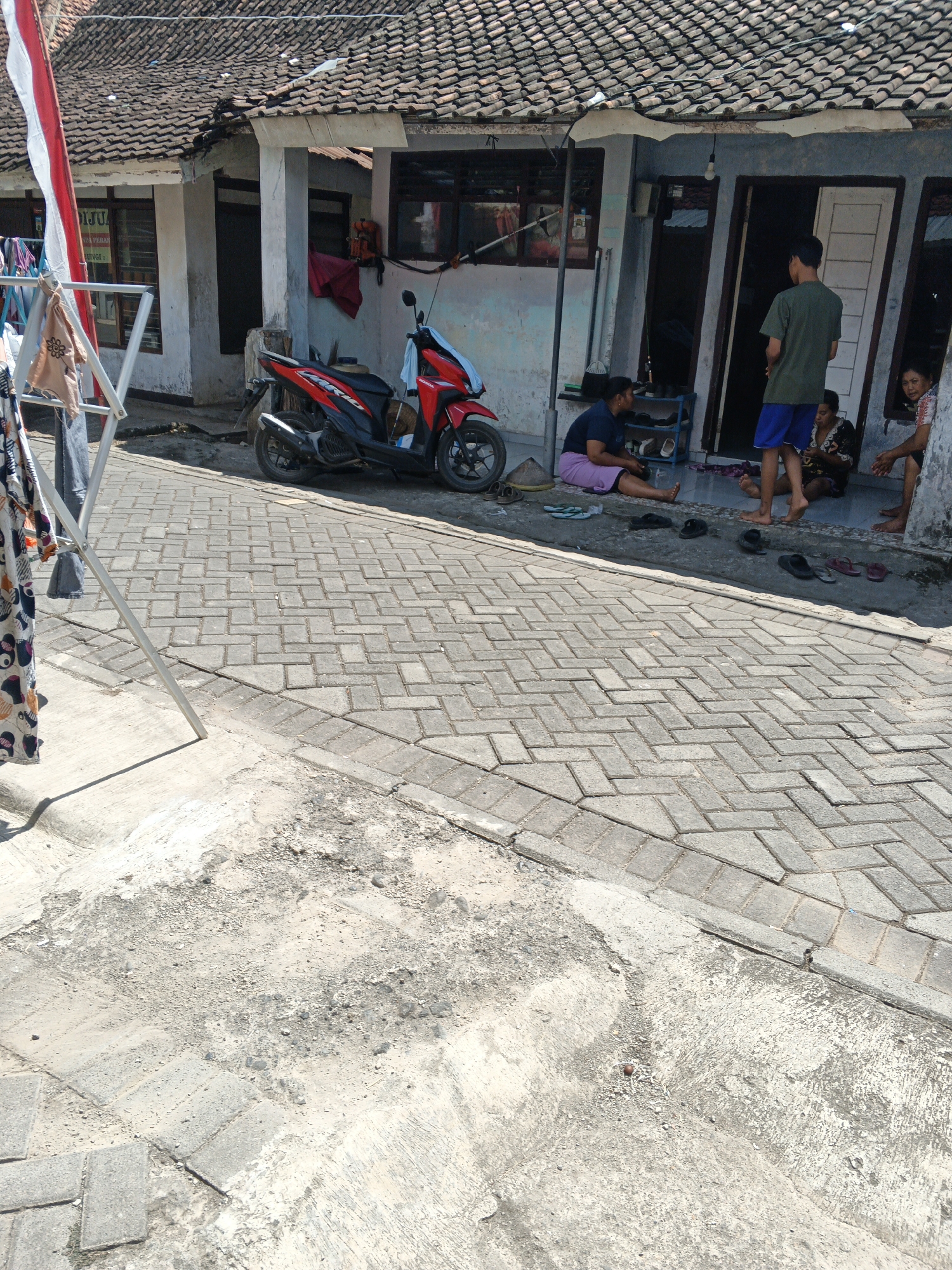 In a residential setting in Singolatren, Indonesia, a red and black motorcycle is parked on a cobblestone pathway. Several individuals are present. A young man wearing a green t-shirt and blue shorts stands near a doorway. To his left, a woman in a dark blue shirt and light purple skirt sits on the ground, facing away from the viewer. Another woman, wearing a dark patterned top, sits on the ground to the right of the young man, looking towards the camera. A third woman, partially visible, is seated to the far right, also looking towards the camera. Laundry is drying on a rack near a building with a tiled roof. The scene appears to be an outdoor common area in front of multiple residences.