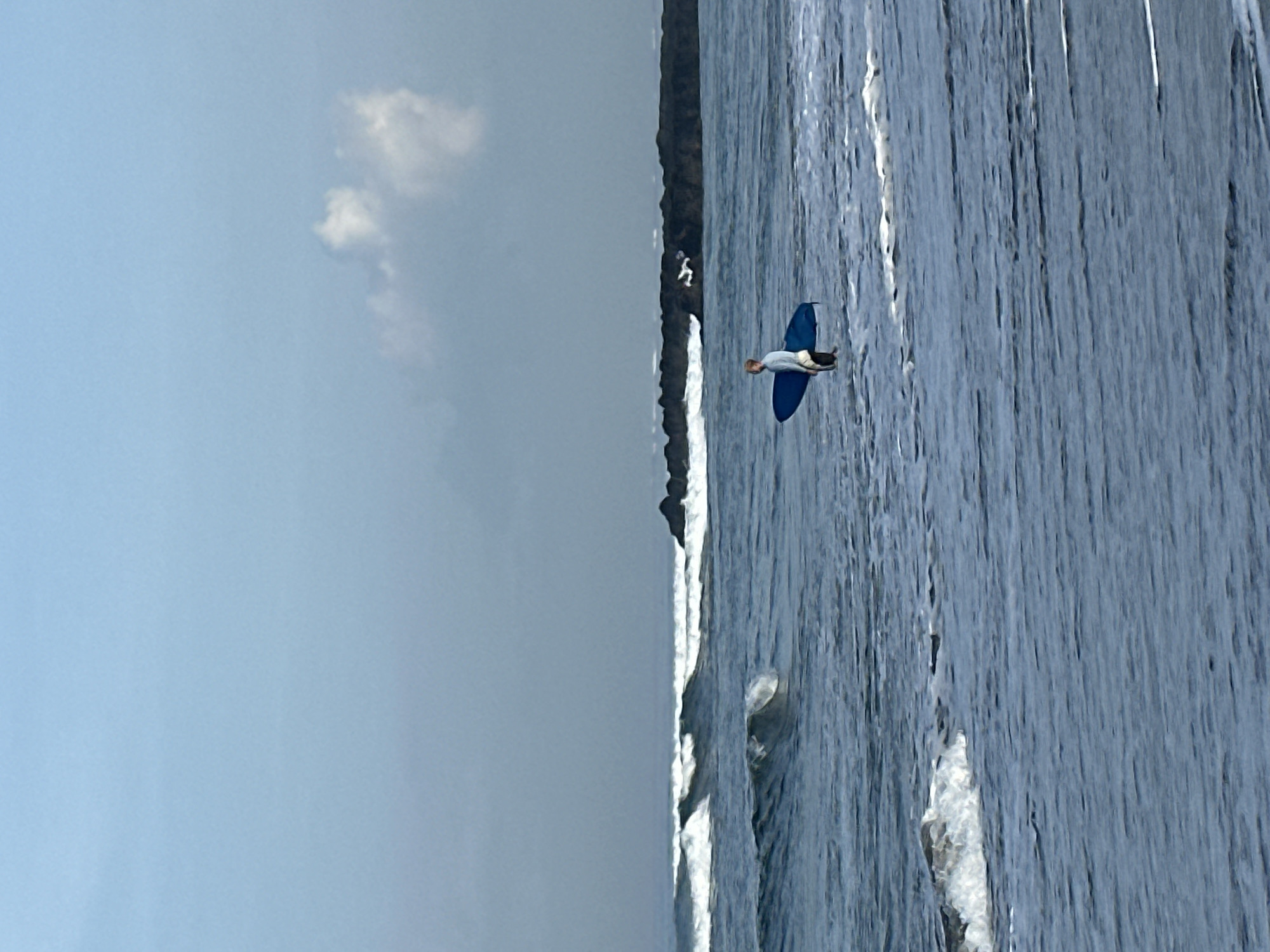 A solitary person is standing in the shallow ocean water, holding a blue surfboard. The individual is wearing a light-colored long-sleeved shirt and dark shorts. The water is relatively calm with small waves breaking. In the background, there are larger waves further out and a rocky formation protruding from the sea. The sky is overcast and a pale blue with a few wispy white clouds. The scene appears to be a beach in North Kuta, Indonesia.