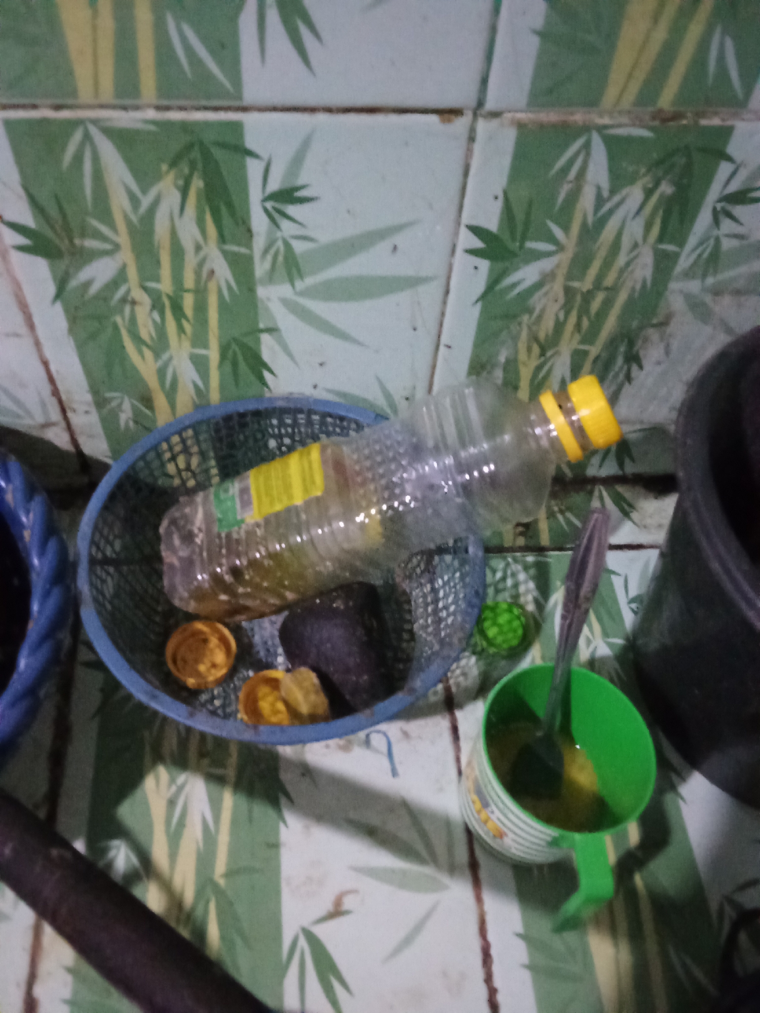 The image depicts a close-up, slightly blurry view of various items arranged on a surface with a green and white bamboo-patterned wallpaper or covering. In the foreground, a dark cylindrical object, possibly a rolling pin or a piece of wood, lies horizontally. To the right, a green mug with a white striped pattern contains a yellow liquid and a spoon. Above and slightly to the left of the mug, a blue woven basket holds a clear plastic bottle with a yellow cap, lying on its side. Within the basket, there are also two small, round, yellow objects and a dark, irregular-shaped object. Partially visible on the left is a section of a blue woven object, possibly another basket or container. The background consists of the bamboo-patterned surface. No people are visible in the image. The scene appears to be an indoor setting, likely a kitchen or a domestic area, given the presence of a bottle, mug, and food-related items. The location is not explicitly identifiable as Pemalang, Indonesia, based solely on the visual information provided.