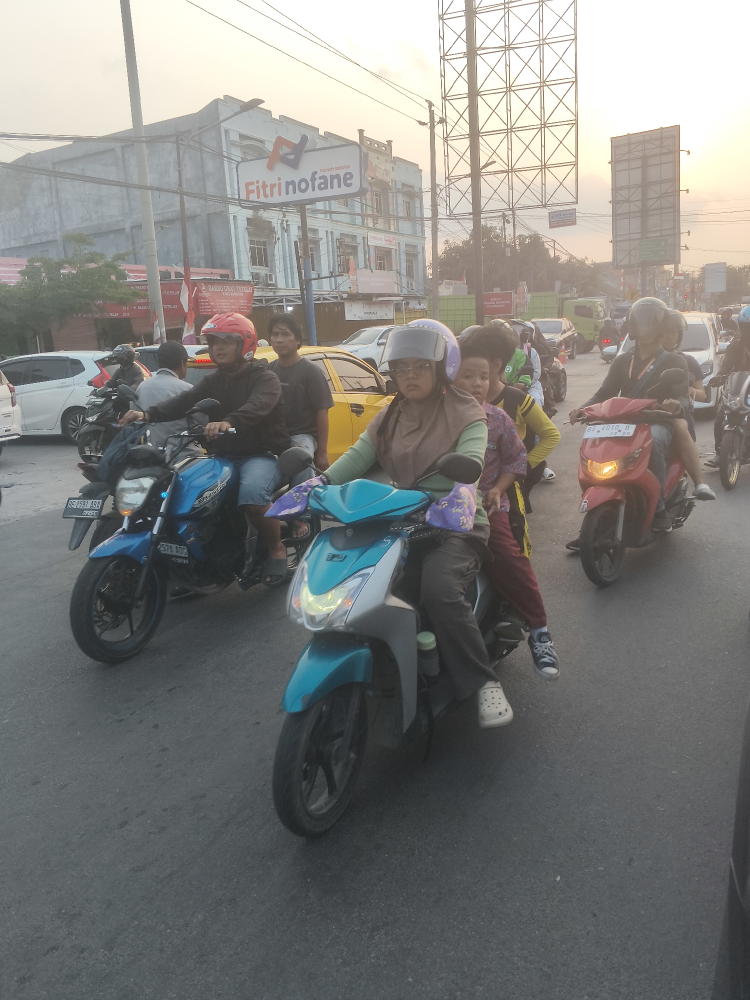 The image depicts a street scene in Bandar Lampung, Indonesia, possibly during dusk. Multiple motorcycles are navigating a busy road, suggesting a traffic jam or congestion. On the foreground, a woman wearing a hijab and glasses is riding a light blue scooter, with a child sitting behind her. Another motorcycle, a blue Yamaha, is to her left, with a man in a red helmet driving and another man behind him. To the right, a red scooter is visible, carrying a man and a child. In the background, buildings line the street, with a prominent "Fitrinofane" sign on one of them. Billboards are also visible, indicating a commercial or advertising area. The overall environment is urban, with the presence of various vehicles and infrastructure.