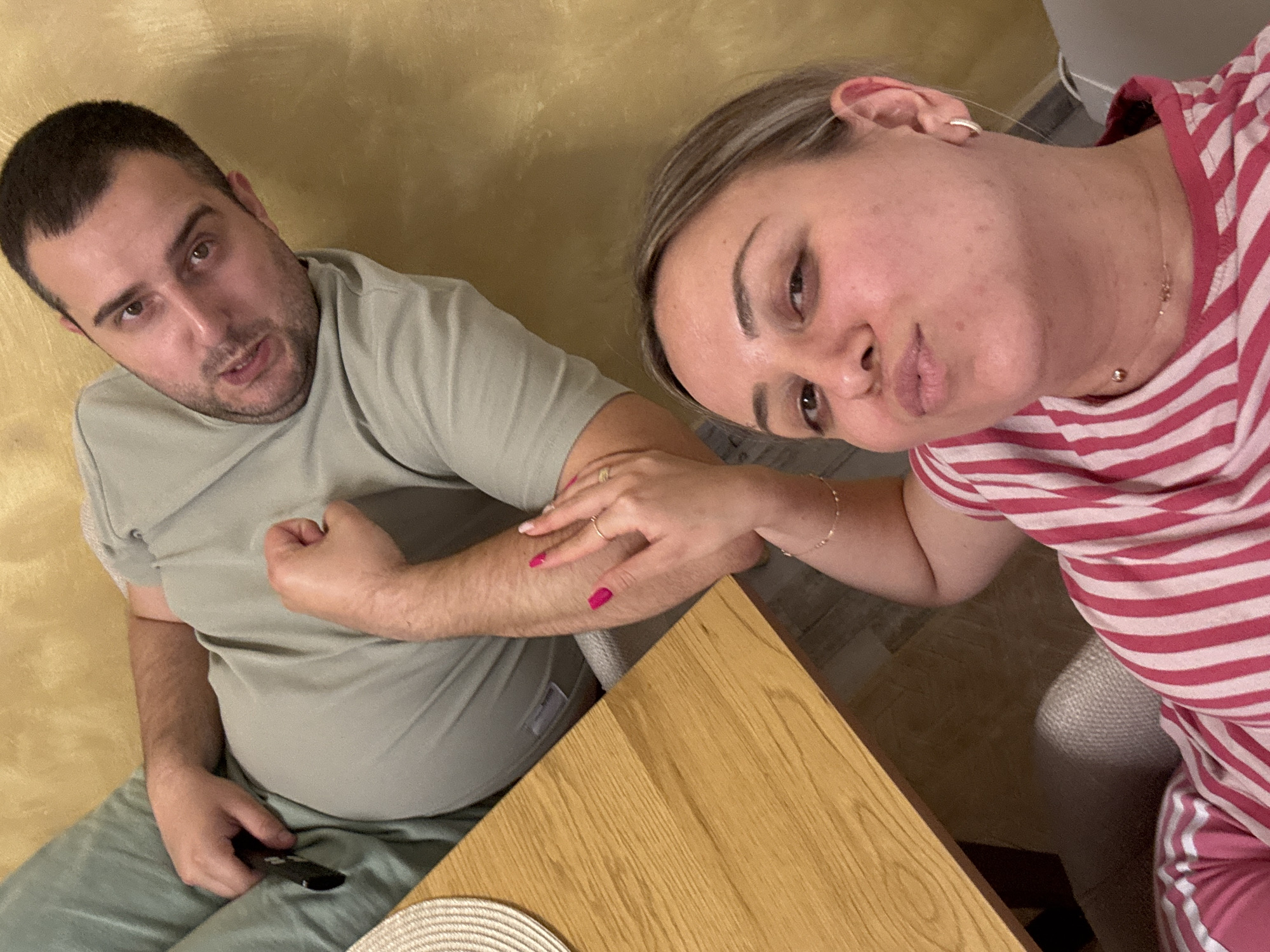 The image shows a man and a woman in an indoor setting. The man is wearing a light green t-shirt and matching pants and is seated with a remote control in his hand. The woman is wearing a pink and white striped shirt and pink pants. She is leaning over the man, resting her hand on his arm, and making a kissing face towards the camera. They are seated at a light brown wooden table on which is a coaster. The background consists of a wall with a mottled yellow and brown texture and an object in white and gray. The location is not apparent from the image.
