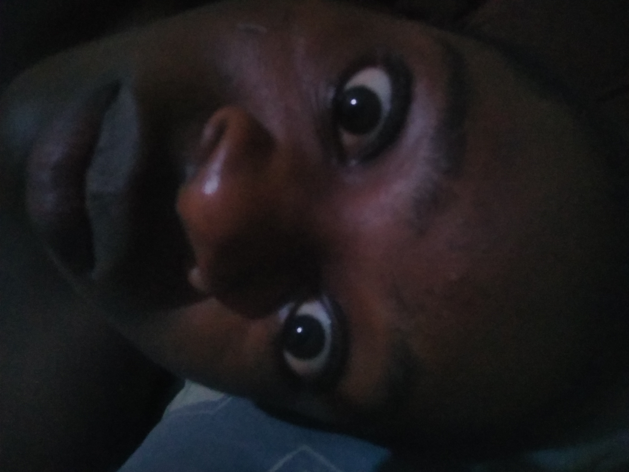 A close-up, slightly angled portrait of a person with dark skin. The person's eyes are wide open, the nose is prominent, and the lips are full. The background is dark and indistinct, but a blue surface is visible at the bottom of the frame. The location of Shangisha, Nigeria, is not apparent from the image.