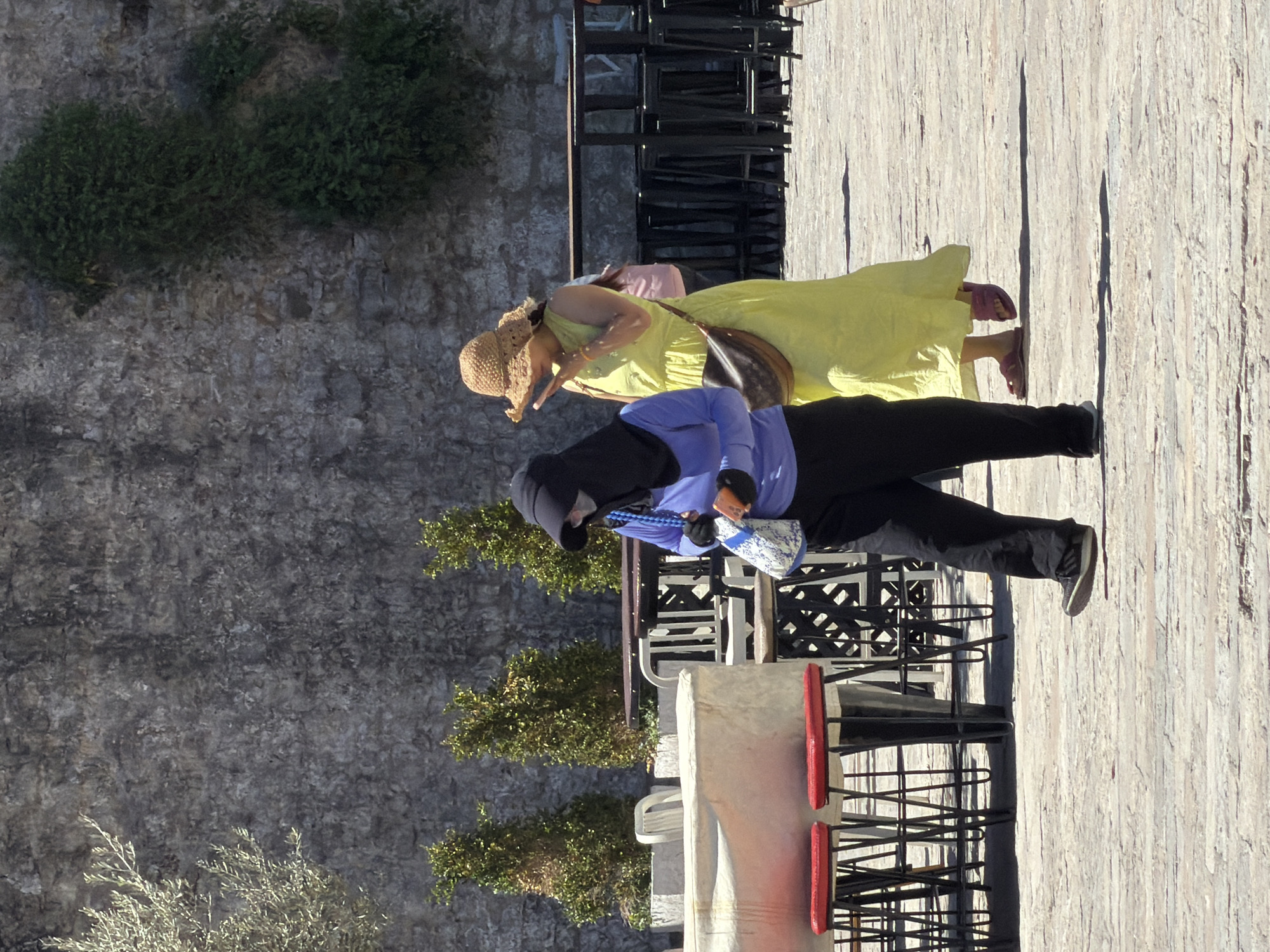 Two women walk along a stone-paved path in Budva, Montenegro, near a stone wall covered in vegetation and a stack of chairs. One woman wears a yellow dress and a straw hat, carrying a brown purse. The other woman wears a blue top, black pants, and a black head covering, carrying a patterned bag.