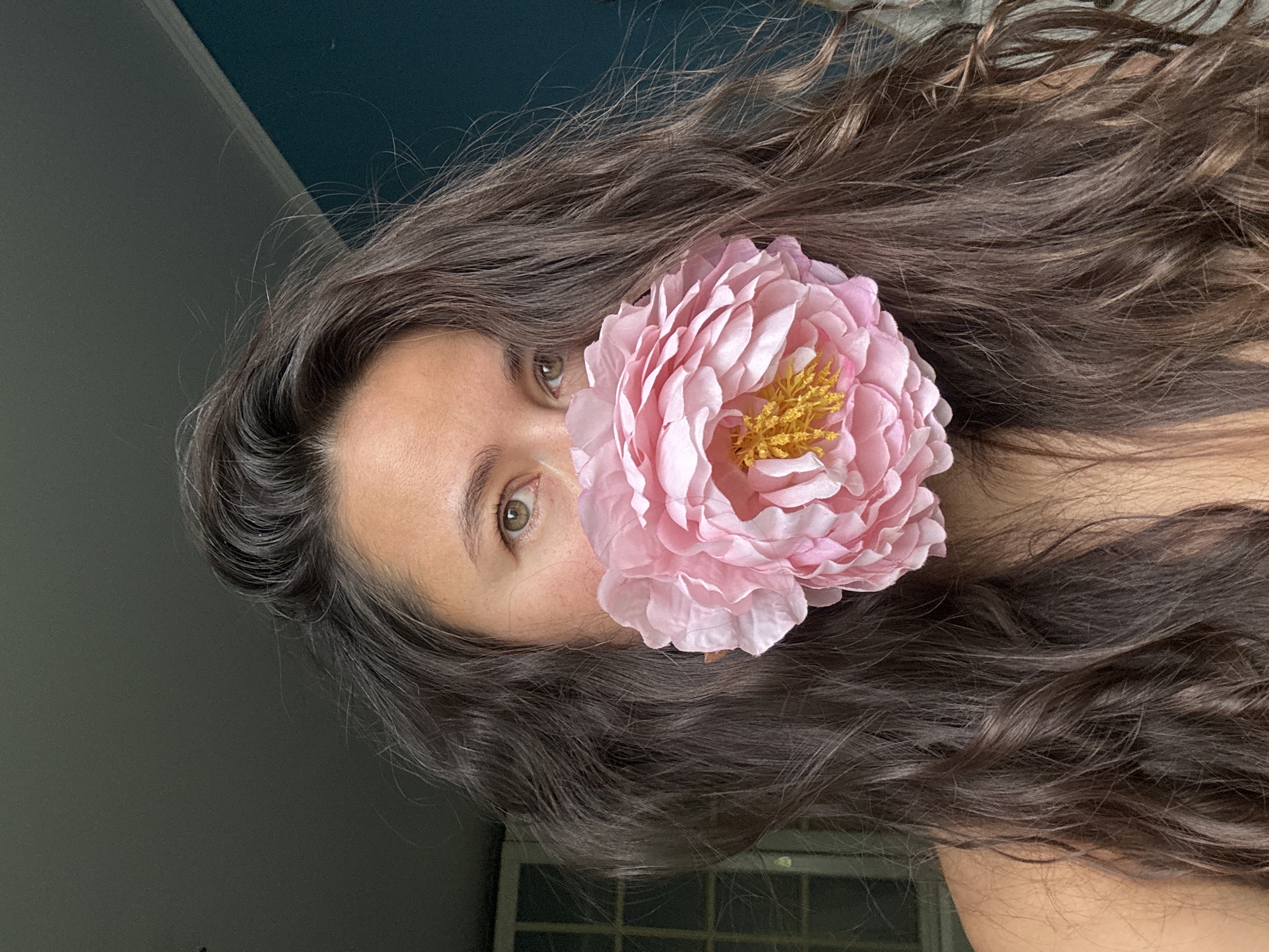 A portrait photograph shows a woman with long, wavy brown hair covering part of her face. The hair is styled loosely around her head and shoulders. A large, light pink artificial flower is positioned over the lower portion of her face, obscuring her mouth and part of her cheek. The flower has densely layered petals and a yellow center. The woman's left eye is visible, showing an iris with shades of green and brown. Her skin appears to be fair. The background is a mix of dark and light gray, possibly walls or an interior space. The location is not evident in the image.
