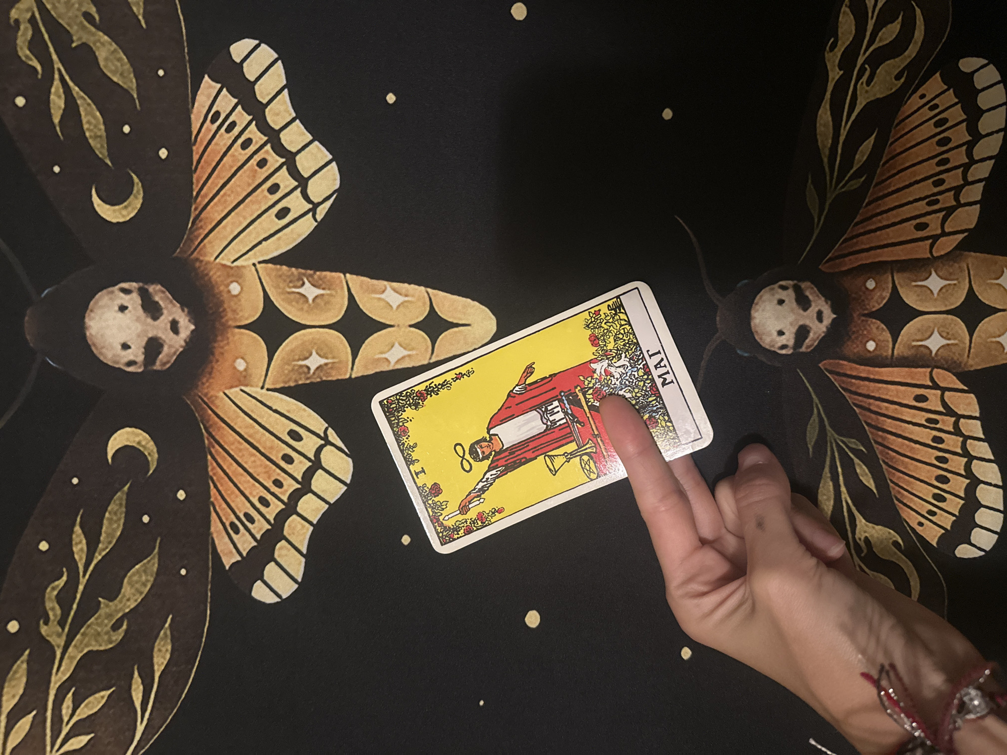 A hand is shown in the lower right of the frame, with two fingers extended, appearing to point at a tarot card. The card, labeled "8 MAI", depicts a figure in red robes holding a wand. The card is placed on a black cloth decorated with large, stylized moth wings in shades of orange and yellow. The wings feature crescent moons, leafy designs, and circular patterns. The scene is lit evenly, suggesting an indoor setting. The overall mood is mystical and focused on the tarot reading.