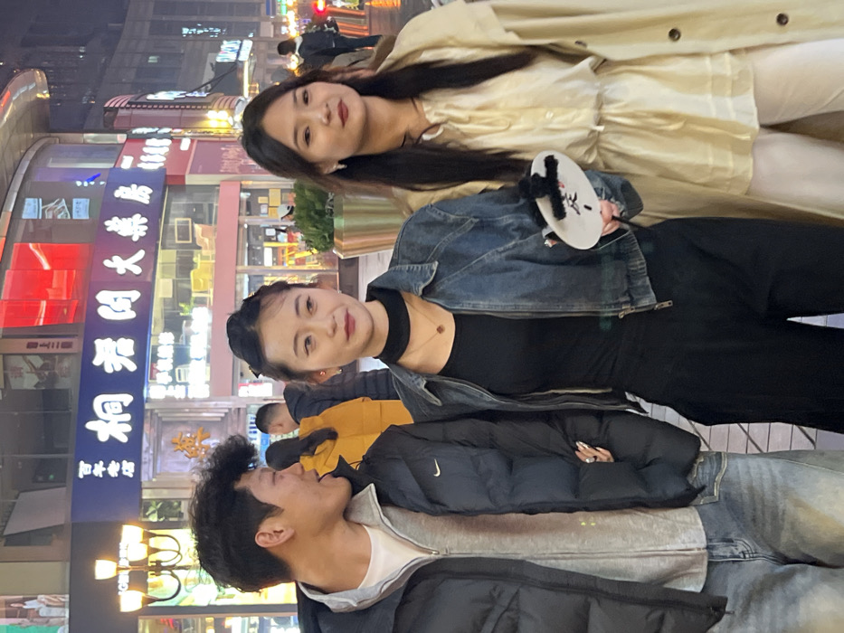 The image depicts three individuals, two female and one male, walking on an illuminated urban street at night in Yuzhong District, China.
The male, positioned on the left, is of Asian ethnicity, with dark, curly hair. He wears a grey hooded sweatshirt under a black puffer jacket with a visible Nike swoosh, and light-wash denim jeans. He is looking to his right, towards the central female, with his mouth slightly open. His right arm is linked with the central female's left arm.
The central female, also of Asian ethnicity, has dark hair styled partly up. She wears a black choker, a gold-colored pendant necklace, a black long-sleeved top, a blue denim jacket, and dark wide-leg pants. She holds a white, oval-shaped object with black text/design and a fluffy black top in her right hand. Her expression is neutral, and she looks directly forward. Her left arm is linked with the male's right arm.
The female on the right, also of Asian ethnicity, has long dark hair. She wears small earrings and a light-colored (cream or pale yellow) long-sleeved tunic with a gathered waist, over light-colored pants. Her expression is neutral, and she looks slightly to her left.
The background features brightly lit storefronts with Chinese characters. A prominent blue sign displays "桐君阁大药房" (Tongjunge Pharmacy) and to its left, "百年老店" (Century-old shop). The environment indicates a bustling commercial area, with other blurred pedestrians and city lights visible in the distance. A golden planter with green foliage is situated behind the central and rightmost individuals.