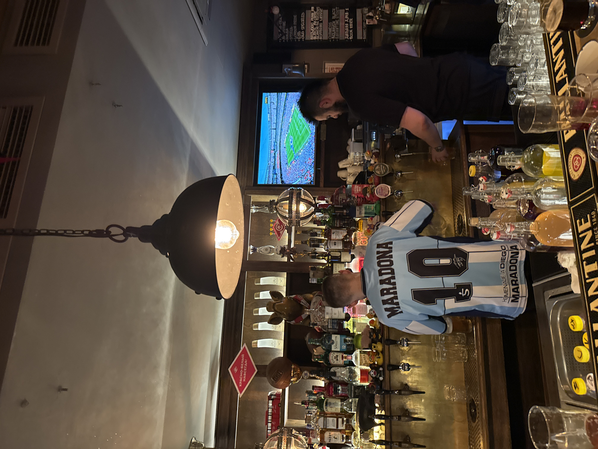 The image depicts the interior of a bar in Moscow, Russia, with a focus on a well-stocked bar counter and two men. In the foreground, a young man wearing a light blue and white soccer jersey with "MARADONA" and the number "10" prominently displayed is standing at the bar. He appears to be holding a glass or a beer tap. Behind him, a bartender with dark hair and a beard, wearing a black t-shirt, is actively working behind the counter, likely serving drinks.
The bar area is illuminated by a large industrial-style pendant light fixture hanging from the ceiling. Shelves behind the bar are filled with various bottles of alcohol, including whiskey, vodka, and other spirits, as well as decorative items such as a soccer ball, a trophy, and a pig figurine. A television screen mounted above the bar displays a live soccer match, indicating a sports-themed environment.
The bar counter itself is made of metal and wood, with multiple beer taps and glasses neatly arranged. In the lower part of the image, a selection of bottles containing colorful liquids, likely liqueurs or mixers, are visible along with stacks of clean glasses. The overall atmosphere suggests a lively and social setting, possibly a pub or a sports bar. The presence of the Maradona jersey and the soccer match on the screen strongly suggests a connection to football culture.