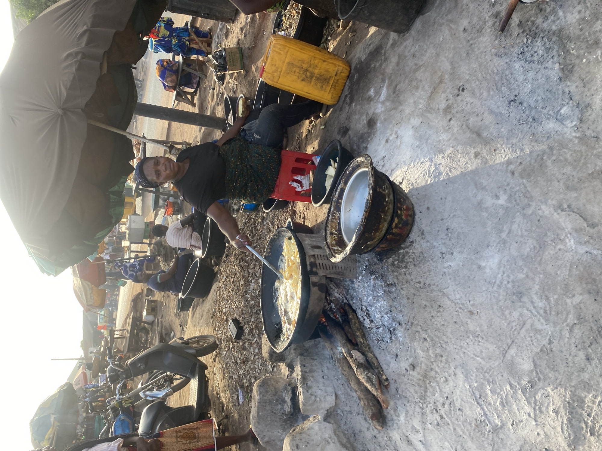 A woman is seated on a red plastic crate on a concrete surface, facing the camera. She is stirring a large, blackened frying pan over an open fire built on top of large rocks. A cooking oil container sits near her on the ground next to another plastic crate. Two large pots rest on a makeshift grill next to the fire. Motorcycles and other individuals are visible in the background, indicating an outdoor, public space. It is likely Rido, Nigeria.