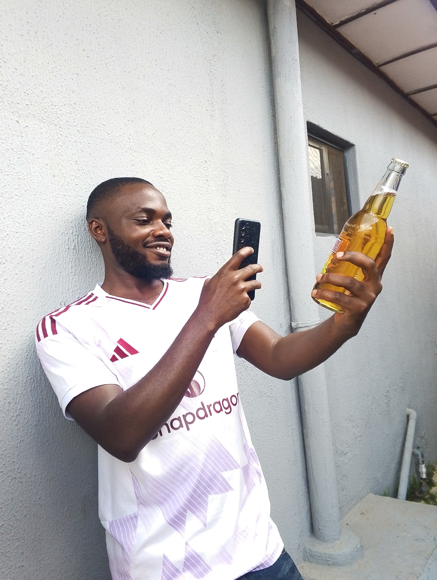 A Black man with a beard and short hair, wearing a white t-shirt with purple stripes and a logo, stands leaning against a light grey textured wall. He is holding a smartphone in his left hand and a bottle of light-colored liquid, possibly beer, in his right hand. He is smiling and looking at the smartphone. In the background, there is a pipe, a window with a dark frame, and another pipe leaning against the wall. The environment appears to be outdoors, possibly in an urban setting. The scene does not provide any specific clues to pinpoint the location to the city of Jos, Nigeria.