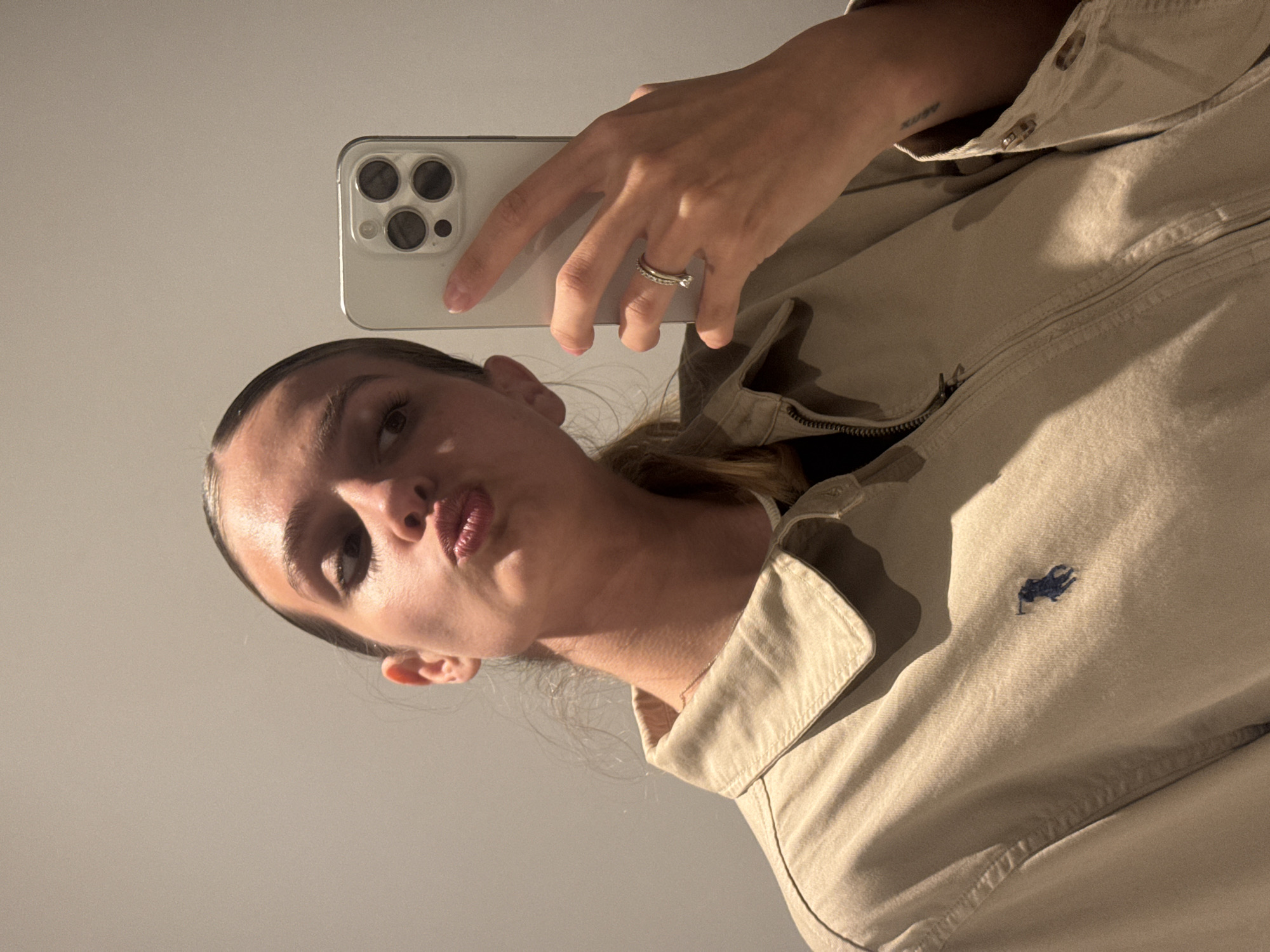 A young adult female is captured in a mirror selfie, with the image rotated 90 degrees clockwise. Her head is positioned towards the bottom-left of the frame, angled upwards, and she is seen from the chest up. She has dark hair styled back and is looking towards the phone with a playful pout.
She wears a light beige or tan collared jacket or shirt. A distinctive small, dark blue embroidered logo of a polo player, indicative of Polo Ralph Lauren, is visible on the right side of her chest. On her left ring finger, she sports two rings, one appearing to be an eternity band. A small, unreadable text tattoo is visible on her inner left forearm.
She is holding a light-colored smartphone, likely an iPhone with a triple-lens camera system, in her right hand to take the photograph. The background is a plain, light-colored surface, suggesting an indoor setting. The warm lighting casts subtle shadows, particularly highlighting her facial features. The image captures a casual self-portrait moment. This photo was taken in Tbilisi, Georgia.