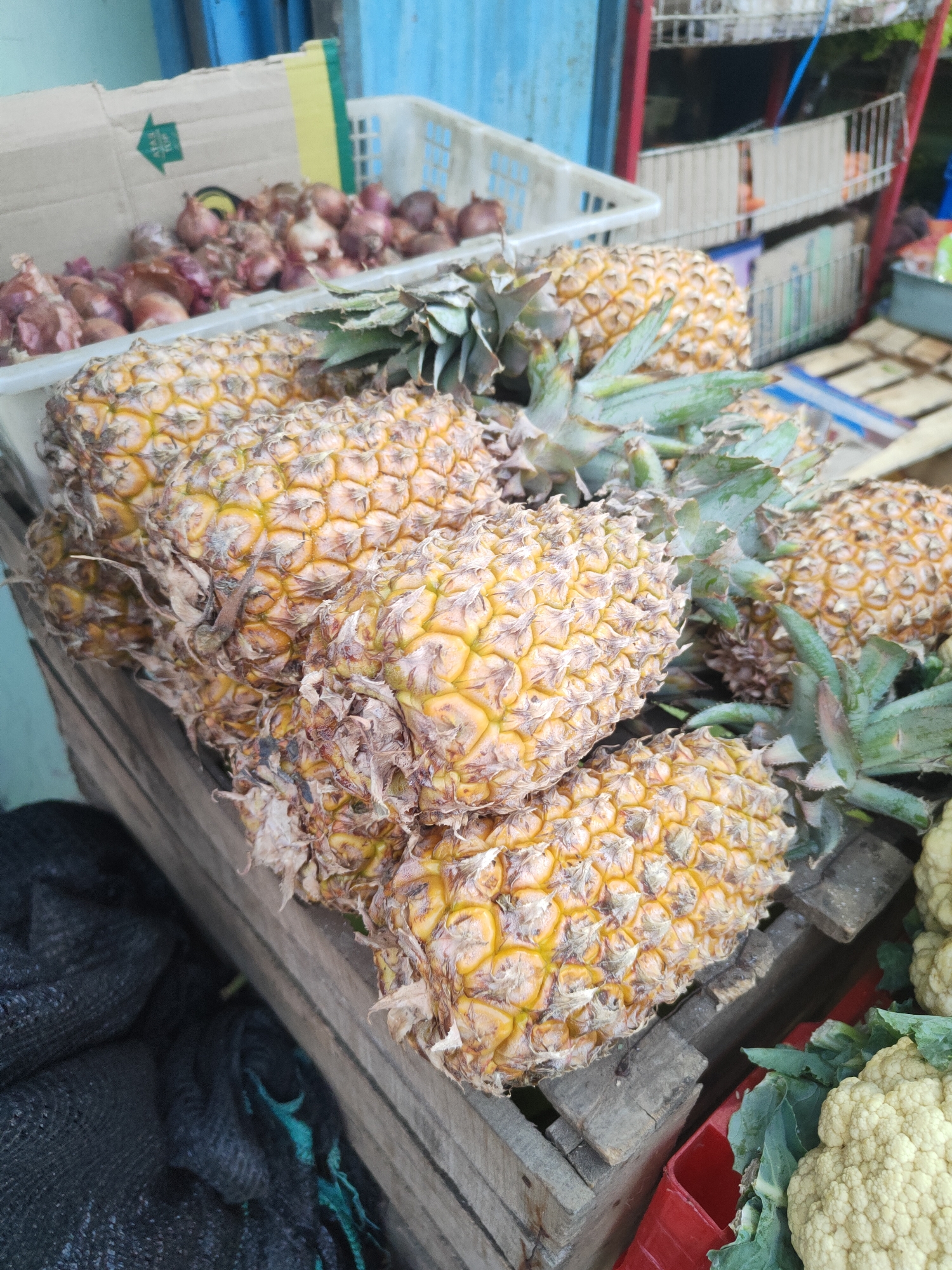 A street vendor's stall in Kauman, Indonesia, displays a pile of ripe pineapples. The pineapples, with their spiky exteriors and leafy green crowns, are a vibrant yellow-orange hue. They rest in a wooden crate, which appears weathered and rustic. Behind the pineapples, a white plastic basket holds a quantity of small, red onions. In the background, shelves filled with various goods are visible, along with blue and red accents suggesting a market setting. To the left of the pineapples, a black mesh bag is partially visible. The overall scene captures a moment of commerce in a local Indonesian market.