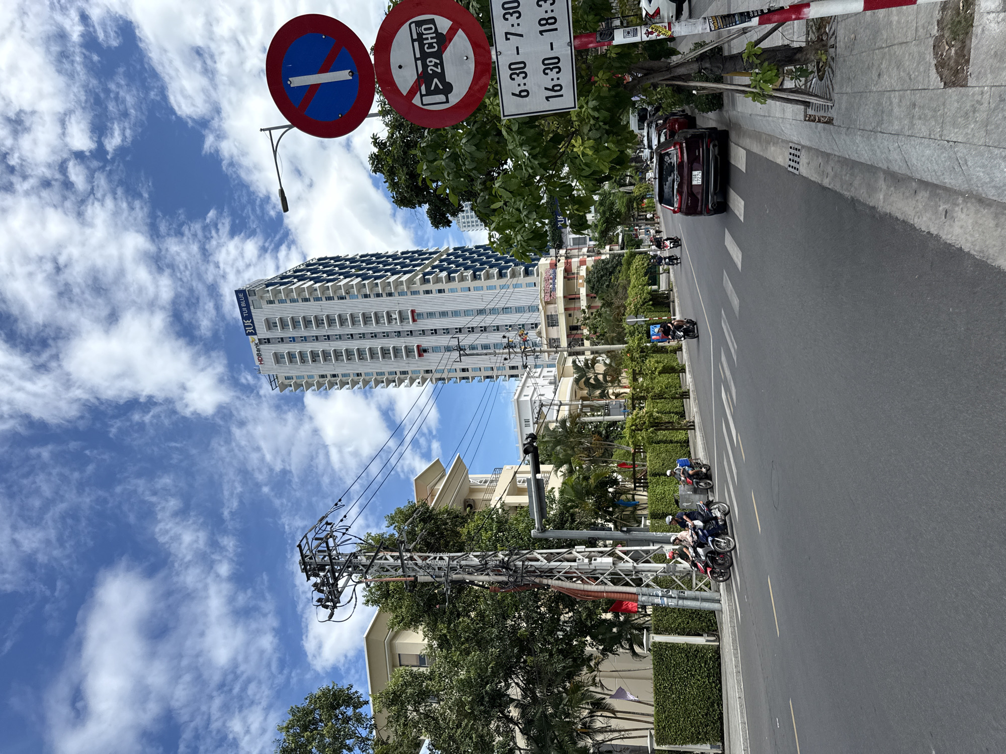 This is a medium shot of a street scene in Nha Trang, Vietnam, on a partly cloudy day. The street is paved with asphalt and features white lane markings and a pedestrian crossing. On the right side of the street, there is a sidewalk with trees, a fire hydrant, and several traffic signs. The traffic signs include a prohibition sign with a red circle and a diagonal red bar over a blue background, indicating "no stopping," and another circular sign with a red border prohibiting buses with more than 29 seats. Below these signs is a rectangular sign with black text on a white background listing times: "6:30 - 7:30" and "16:30 - 18:30."
In the foreground, there are several motorcycles with riders. A dark red SUV is parked on the right side of the road. In the background, there are several buildings, including a tall, modern high-rise building with a blue-tinted facade and white balconies, featuring the text "HPBank" and "TUI BLUE" at the top. Other buildings include a light-colored multi-story building with balconies and trees in front of it. There are also utility poles with numerous wires. The sky is blue with scattered white clouds.
