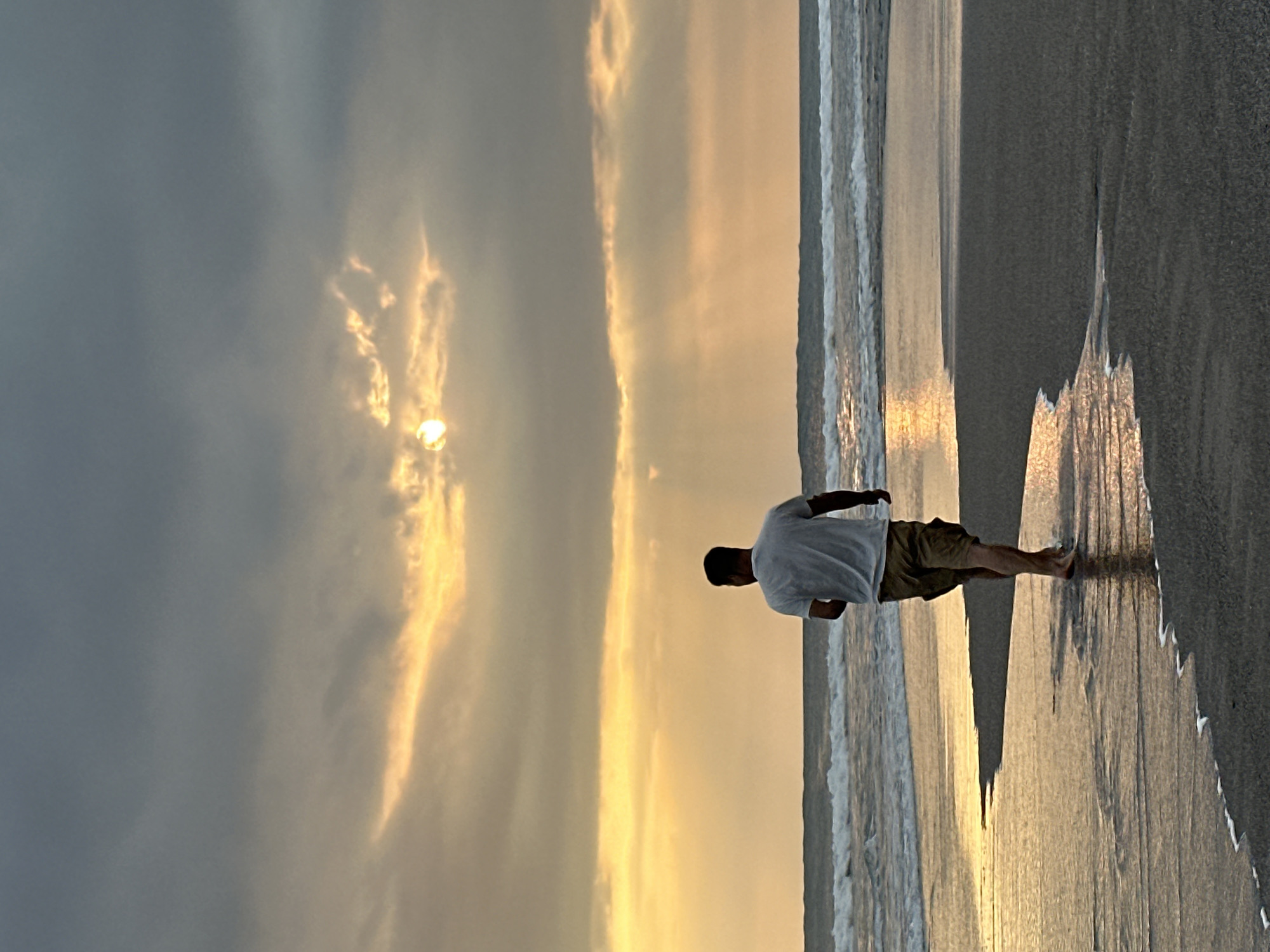 A man wearing a white t-shirt and khaki shorts is walking on a beach in North Kuta, Indonesia, towards the ocean. The sun is low in the sky, casting a warm glow on the clouds and water, creating a dramatic sunset scene. The waves are gently rolling in, and the wet sand reflects the golden light. The man appears to be alone, enjoying the peaceful atmosphere of the beach.