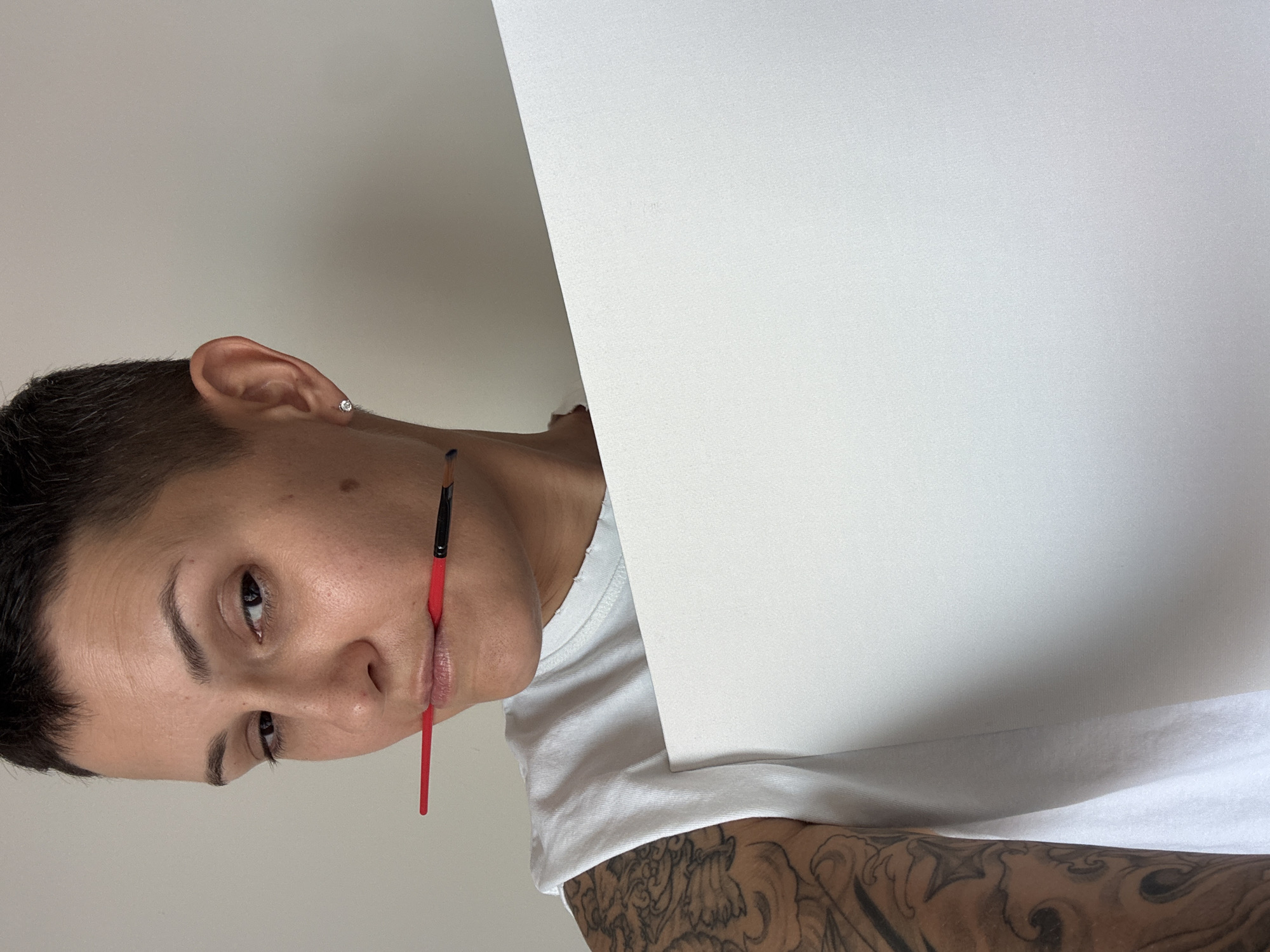 The image shows a person with short, dark hair and a tattoo on their left arm. They are wearing a white, sleeveless shirt and holding a white piece of paper. A red-handled paintbrush is held between their teeth. The person is looking at the camera with their head tilted slightly to the left. The background is a plain, light-colored wall. The image appears to be taken indoors with artificial lighting.
