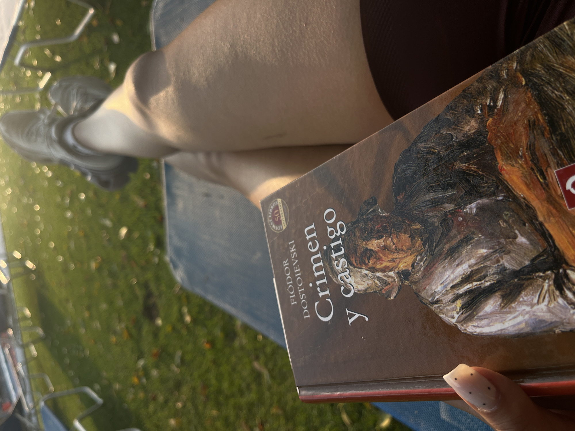 The image shows a person sitting outdoors, with their legs crossed, reading a book titled "Crimen y Castigo" by Fiódor Dostoievski. The person is wearing what appears to be athletic shoes and a dark-colored skirt or shorts. They are outside, on what looks like a grassy area, with some blurred objects in the background. The location is not discernible from the image.