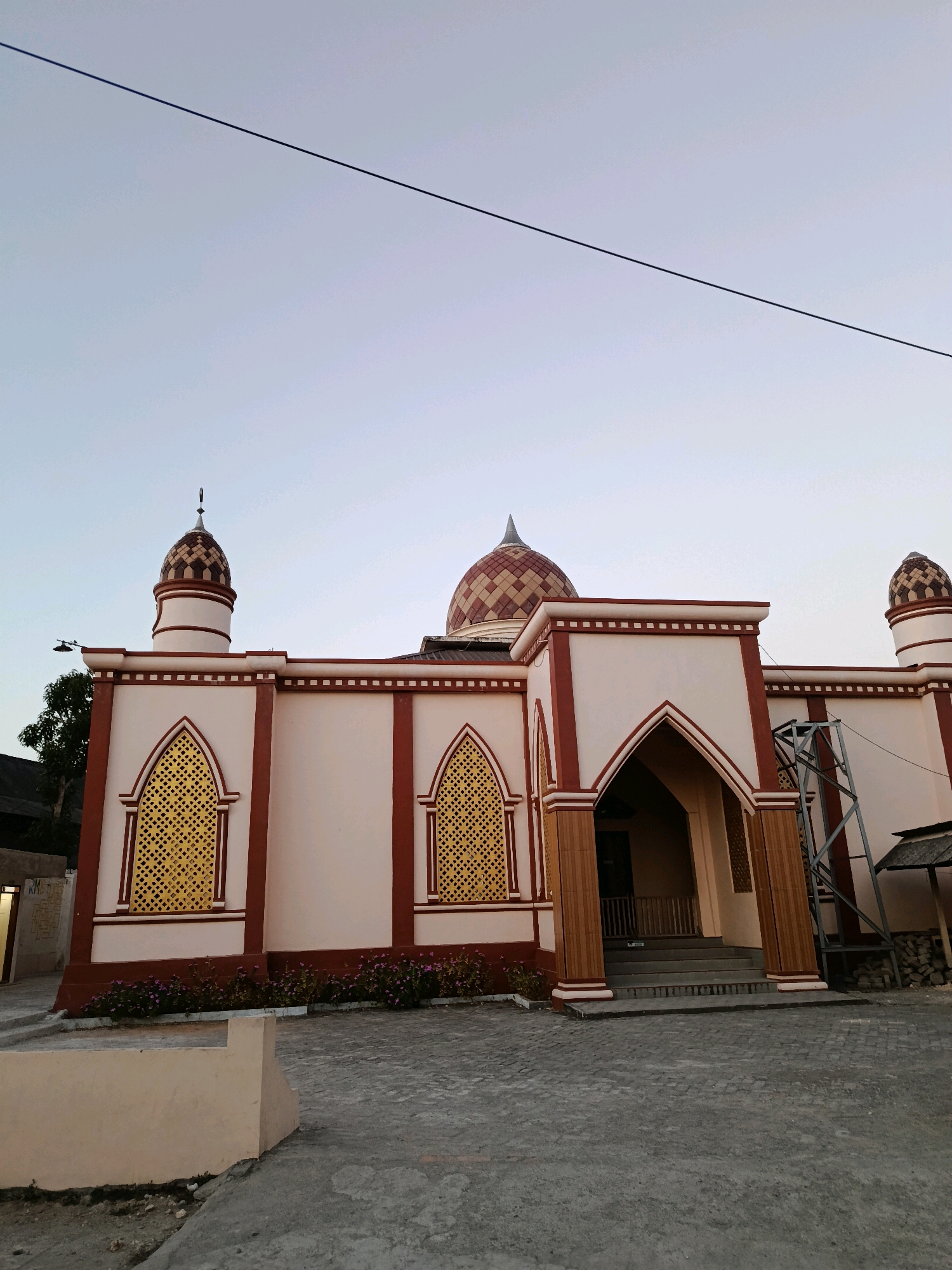 This is a frontal, eye-level, outdoor, full shot of a mosque located in Bulukumba, Indonesia. The mosque has a main dome and two smaller domes atop minarets. The building is painted cream with maroon accents, and features arched windows with golden lattice work. In front of the mosque, a paved courtyard leads up to a set of stairs and an arched entryway. A line of small, pink flowers runs along the base of the mosque. The sky is clear and light blue, with a single dark power line cutting across the upper portion of the frame. The overall impression is one of a peaceful, religious structure in a rural or suburban setting.