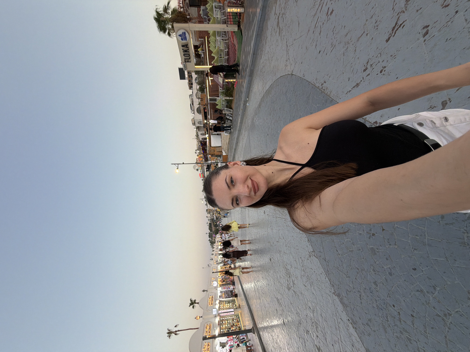A woman with long brown hair and fair skin is taking a selfie outdoors in an urban environment. She is wearing a black one-shoulder top and white shorts with a black belt. In the background, there is a street lined with shops and restaurants, and palm trees are visible. People are walking along the street, and some are seated at outdoor dining areas. The sky is clear and light blue, suggesting it is either early morning or late afternoon. A sign for "FLOKA" restaurant is prominently displayed on one of the buildings. The pavement in the foreground has a distinctive grey, textured pattern. The overall scene appears to be a pedestrian walkway in a resort town.