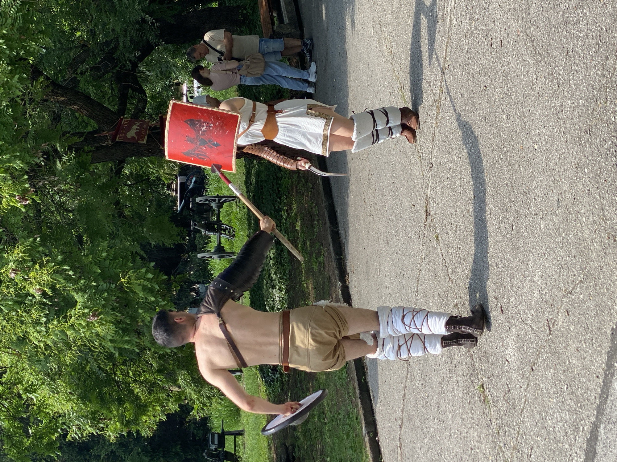 The image captures a historical reenactment event taking place on a paved surface within a park in Pleven, Bulgaria. Two individuals, dressed in ancient Roman gladiator costumes, are positioned centrally, appearing to engage in a simulated combat or demonstration.
The male gladiator, on the left, has his back to the viewer. He wears a bare torso with a brown leather strap across his right shoulder, a black arm guard on his left arm, light brown baggy shorts, and white leg wraps secured with brown lacing over dark boots. He holds a small, round, white shield in his right hand and a wooden stick in his left hand, directed towards the other participant.
The female gladiator, on the right, faces the male gladiator. She wears a white tunic-style dress with a brown belt, a segmented brown arm guard on her right arm, white fur-lined leg coverings, and brown shoes. She holds a large, red, rectangular shield with a black double-headed eagle design, obscuring her face, and a curved blade (sica) in her left hand, pointed towards her opponent.
In the background, to the right of the female gladiator, two spectators in modern attire (one male, one female) are observing the event. The environment is dominated by dense green trees. Historical artillery pieces, resembling cannons, are partially visible in the far left and center-left background. A red banner displaying "LEG. IIII" (Legio IIII Flavia Felix) with a lion emblem is visible hanging from a tree branch behind the female gladiator, reinforcing the Roman theme. The ground is a grey paved surface with minor cracks.