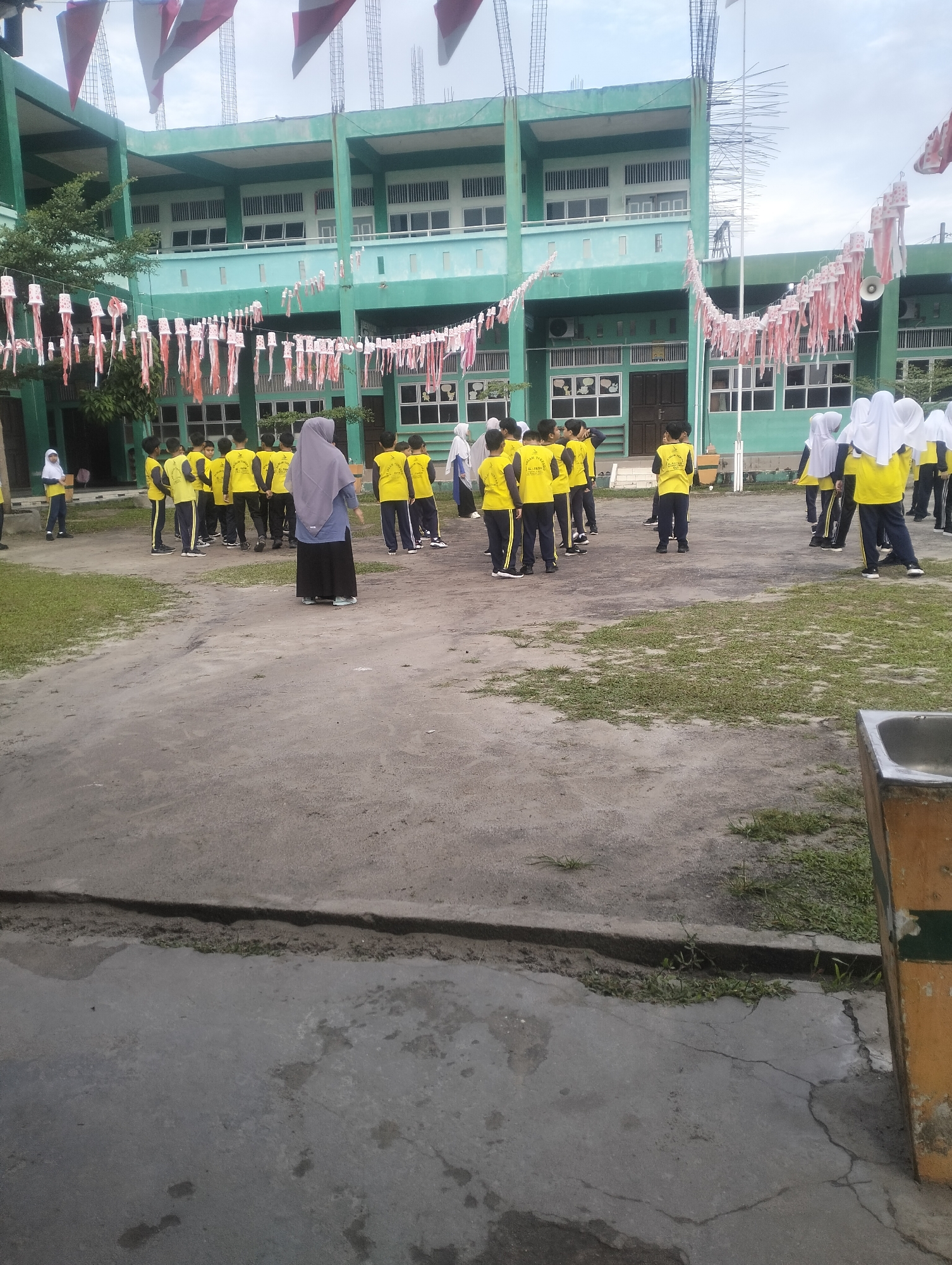 A group of students, some wearing yellow shirts and dark pants, are gathered in an outdoor courtyard of a green, multi-story school building. Some students are wearing headscarves. A woman in a grey headscarf and dark skirt stands in the foreground, facing away from the camera. Decorations, including what appear to be paper streamers and flags, are strung across the courtyard and draped along the building. The ground is a mixture of dirt and patchy grass. The building has multiple windows and balconies. In the background, more students are visible, as are parts of the building's structure, including scaffolding. To the right of the image, a portion of a yellow and green structure, possibly a trash receptacle or monument, is visible. The sky is overcast. The location is identified as Air Jamban, Indonesia.