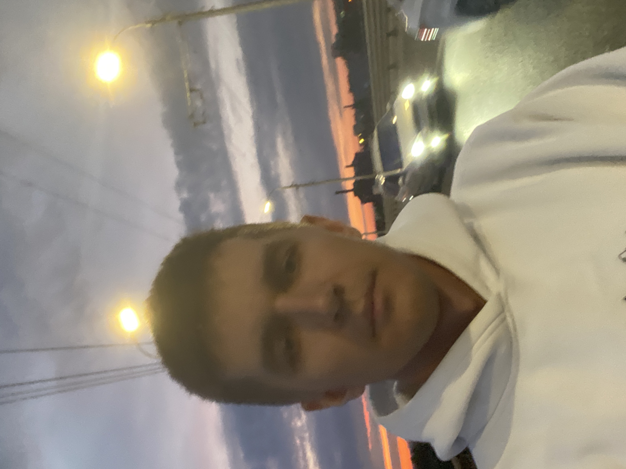 The image shows a young Caucasian male taking a selfie outdoors at dusk. He is wearing a white hoodie. In the background, there are streetlights, power lines, and cars on a wet road. The sky is cloudy with hints of orange and pink from the sunset. The setting appears to be a bridge or overpass in the city of Novokuybyshevsk, Russia, indicated by the presence of typical urban infrastructure like streetlights and road traffic.