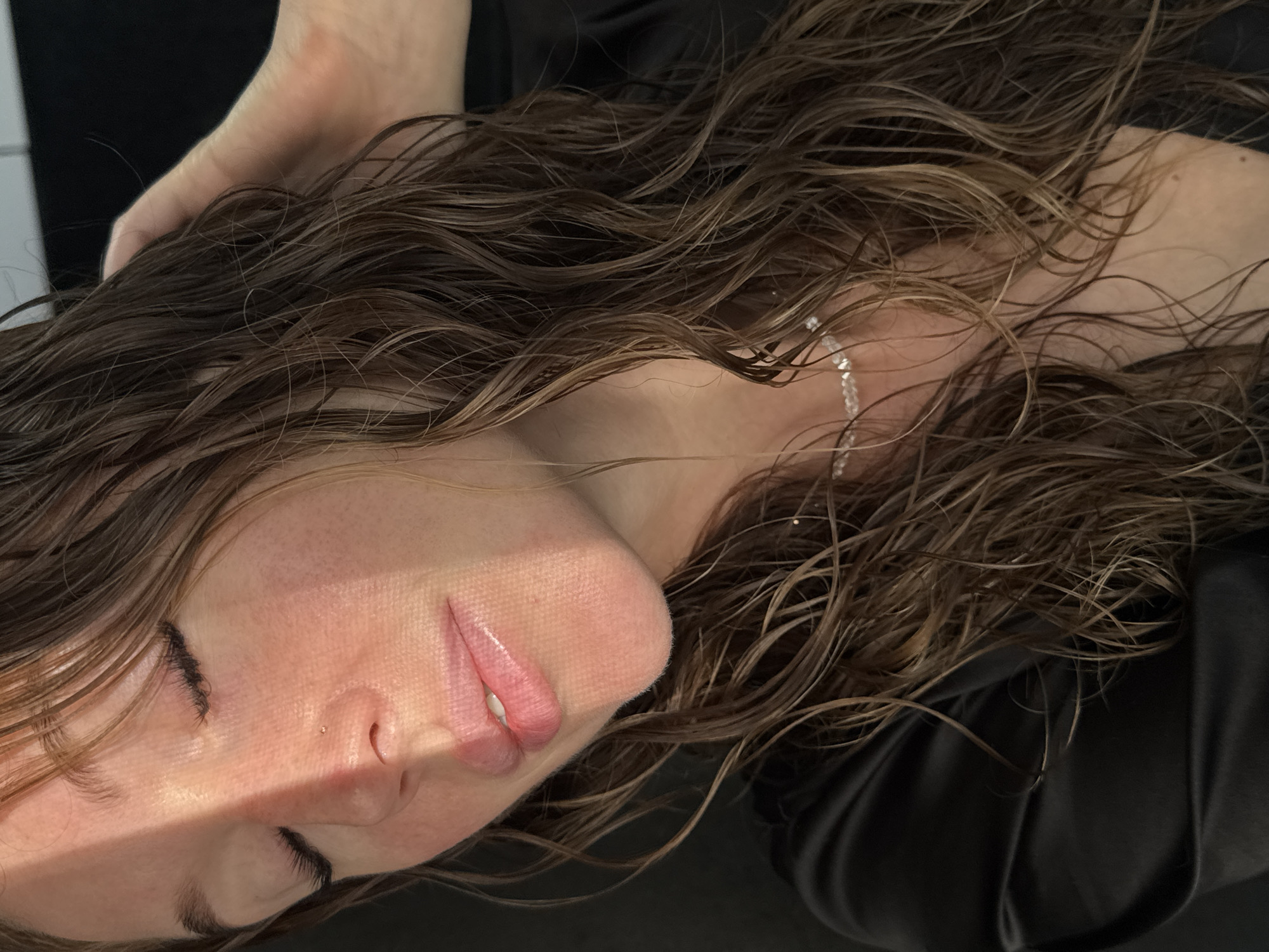 A close-up, tilted shot features a person's face and hair. The person is lying down with their eyes closed. The skin is fair, and the lips are slightly parted. Light illuminates the right side of their face, creating a distinct shadow. Their hair is long, wavy, and appears wet, with strands scattered around the face and neck. A sparkling bracelet is visible on the wrist or arm. A hand is seen touching the top of the head. The person is wearing a dark-colored garment. The background is partially visible and includes a white wall. The image does not provide any location in the city of Chaikovsky, Russia.