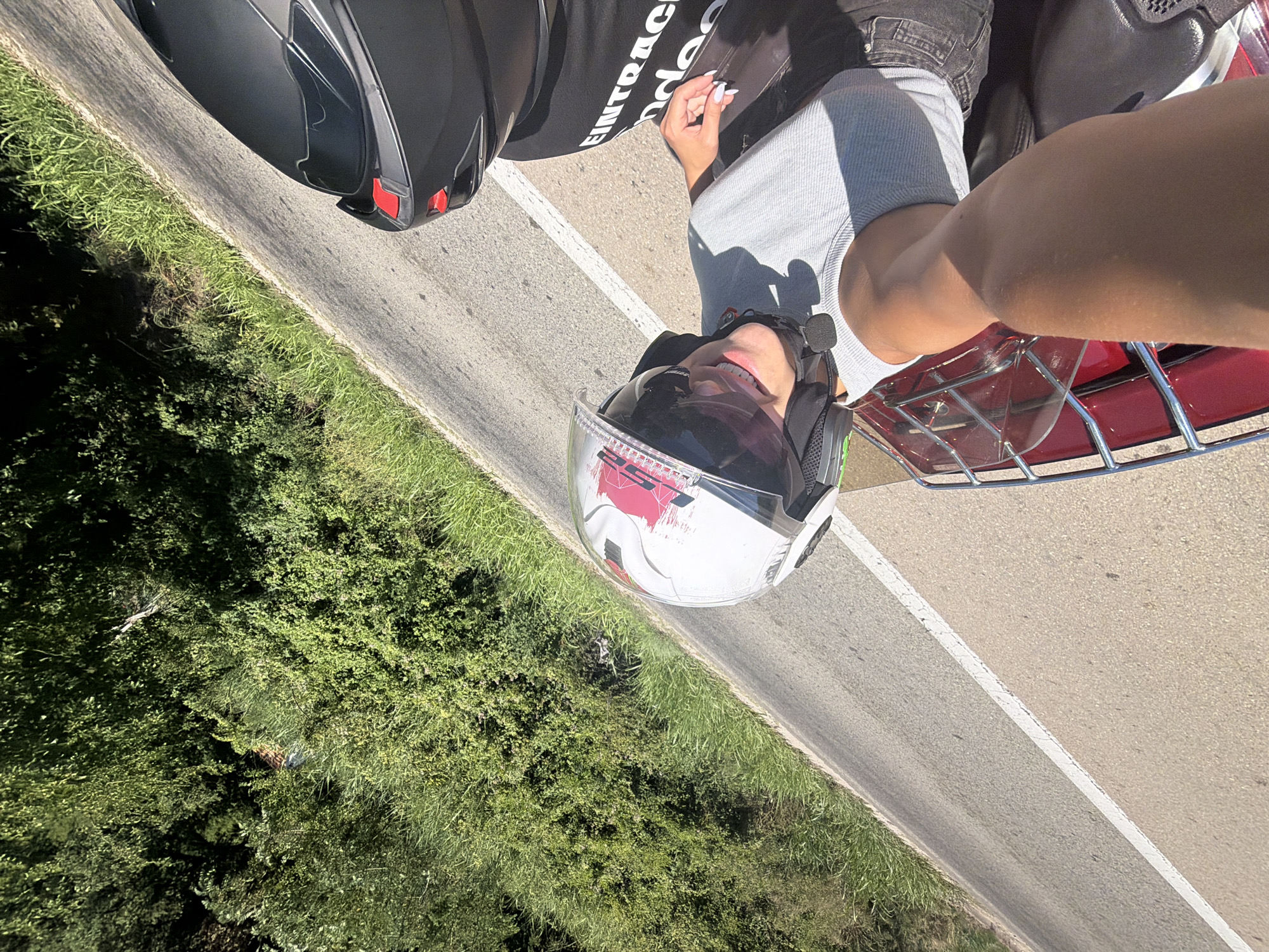 In Mišići, Montenegro, on a sunny day, two individuals are riding on a motorcycle. The person in the foreground, wearing a white helmet with red accents and a grey tank top, is smiling and looking towards the camera. They have fair skin and white painted fingernails. The second person, positioned behind the first, is wearing a black helmet and a black t-shirt with white text that appears to read "EINTRACHT". The motorcycle has a red luggage rack. The scene is set on a paved road with a white line marking the lane, bordered by lush green vegetation and trees on the left side of the road. The perspective is from a high angle, looking down at the riders.