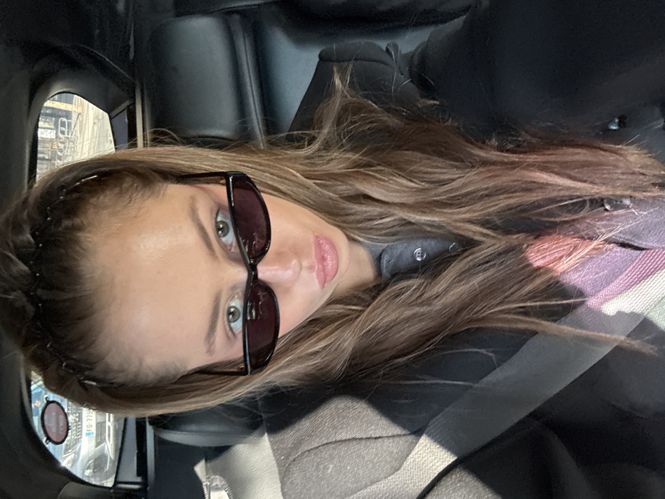 A female individual is seated in the passenger rear seat of a vehicle, likely in the city of Unknown, Unknown. She has long, light brown hair, partially secured by dark hair clips on top, and wears dark, rectangular sunglasses, revealing light-colored eyes. Her lips are pursed. She is dressed in a dark blazer or jacket over a grey collared shirt, with a multi-striped scarf (pink, purple, green) visible lower on her torso. A light-colored seatbelt crosses her left shoulder and chest. The interior of the car features dark upholstery. Through the rear window, a light-colored car with a partially visible European license plate (FG 779xx) is seen, indicating an outdoor, urban environment during daylight. The individual is positioned as if taking a selfie, with no other people present or interacting within the frame.