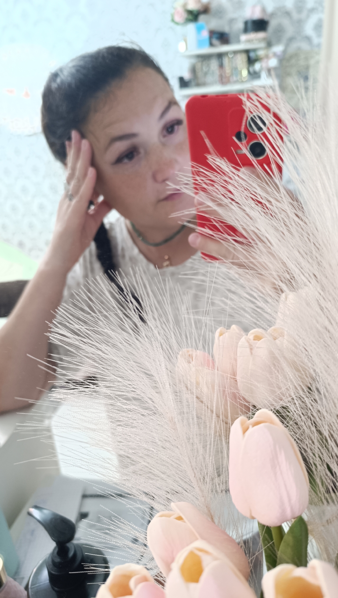 A young woman is reflected in a mirror, holding a red smartphone. She is looking at the phone, with her left hand touching her forehead. She has her hair pulled back and is wearing a light-colored top with a beaded necklace. In the foreground, a bouquet of pale pink tulips and fluffy white reeds are visible, also reflected in the mirror. The background of the reflection shows shelves with various items. This scene appears to be in a home or a salon, possibly in the city of Chaikovsky, Russia, given the provided information.