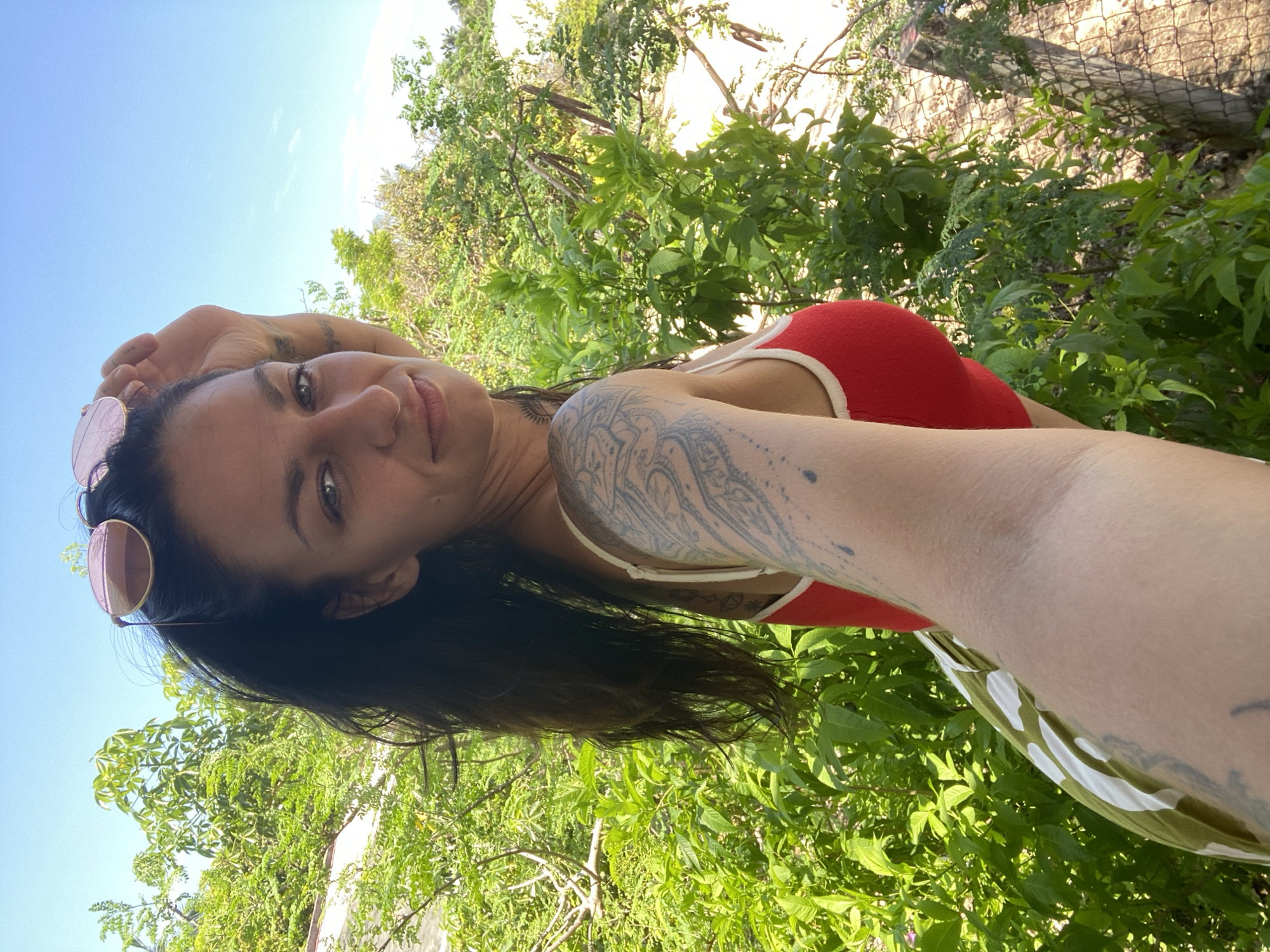 A woman is taking a selfie outdoors in a natural setting. She is lying down with her head tilted towards the camera, and her arm is raised, holding the phone. She is wearing a red halter-neck swimsuit top and sunglasses with pink lenses. Her hair is dark and falls over her shoulder. She has tattoos on her arm and shoulder. The background is lush with green foliage, suggesting a tropical or garden environment. The sky is visible and appears clear and blue, indicating it is daytime with fair weather. The orientation of the image suggests the photographer might be lying on the ground or leaning back. There is no visible text in the image. The scene is casual and relaxed.